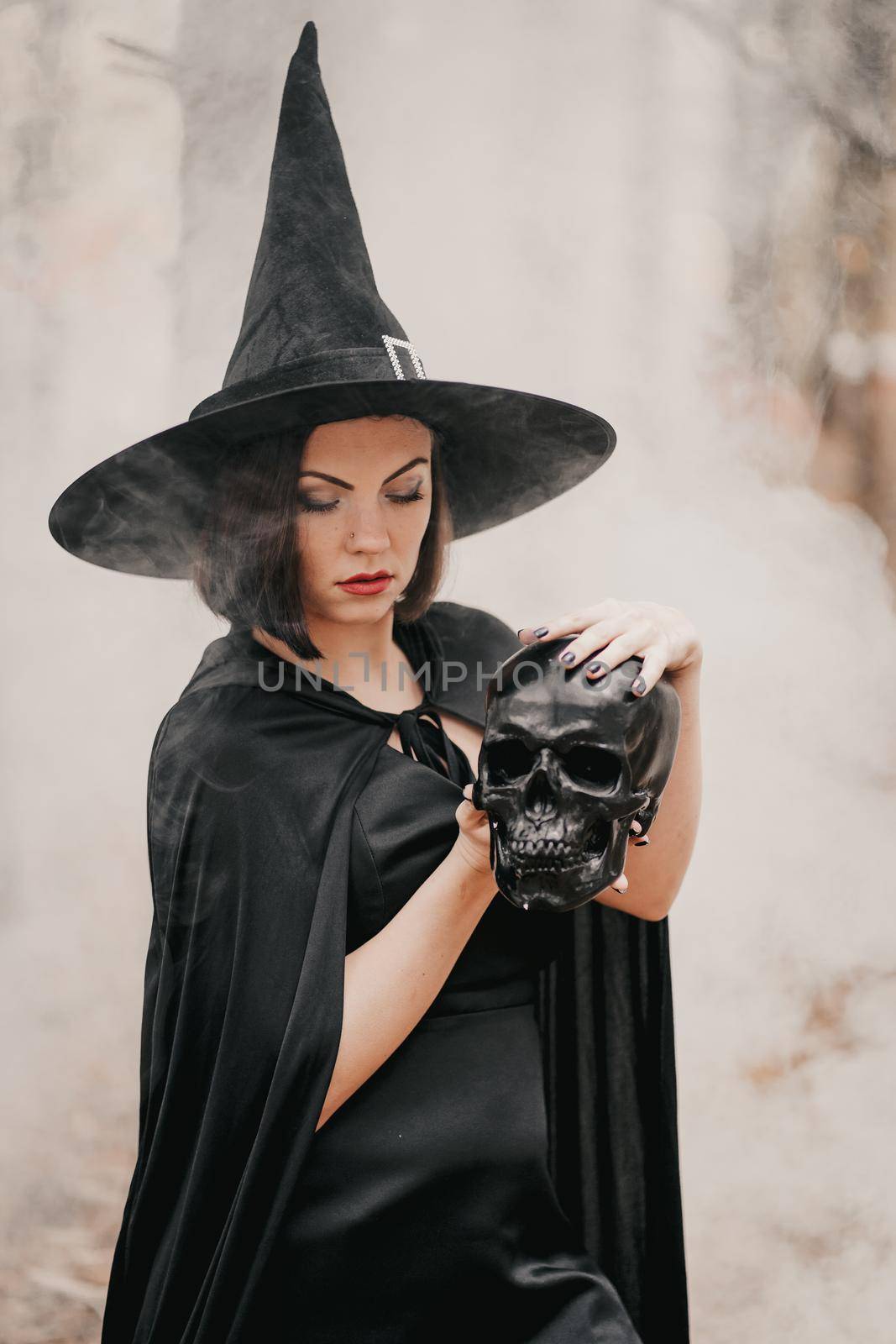 Black witch holding skull in hands, stroking it on autumn forest background. Horror, halloween, cosplay holiday, magic concept. High quality photo