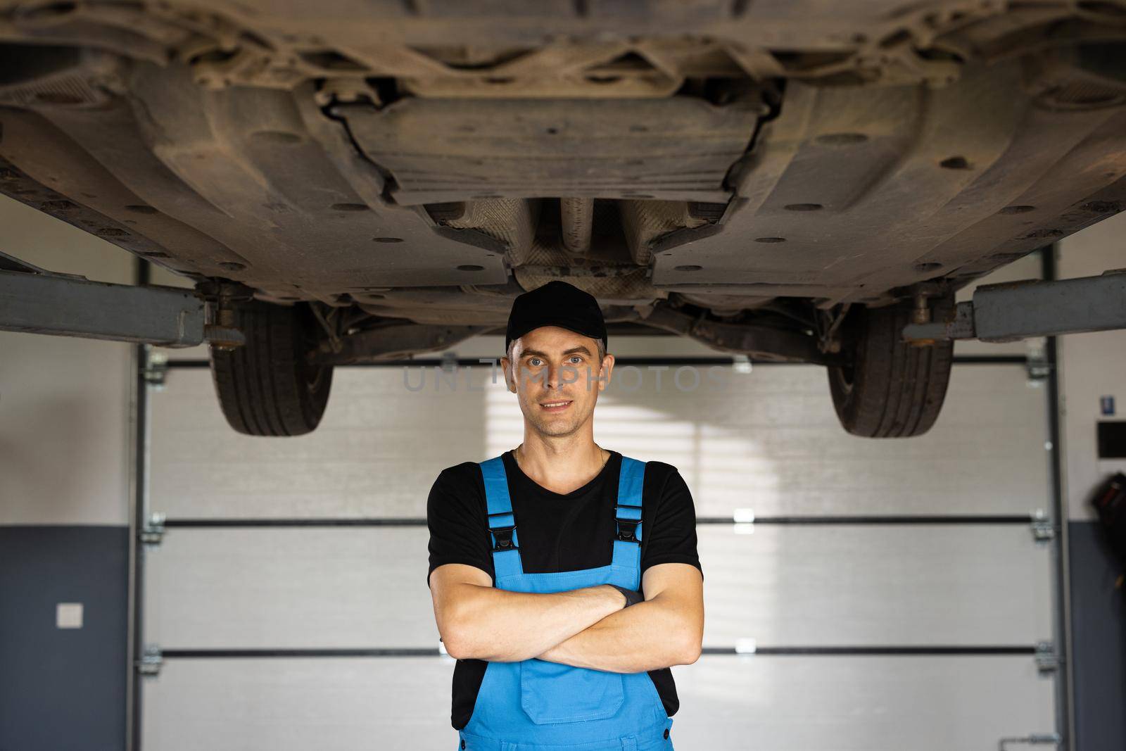 Mechanic man standing in front of the garage. Joyful young bearded man motor mechanic in overalls standing in workshop. Motor mechanic. Concept of small business, own business by uflypro