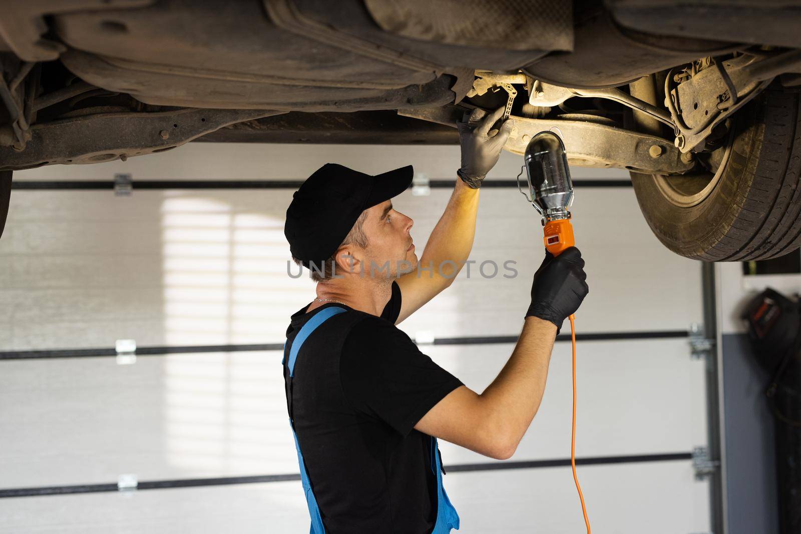 Professional mechanic is working on a car in a car service. Auto mechanic man in blue overalls with lamp working at workshop. Car service, repair, maintenance and people concept.