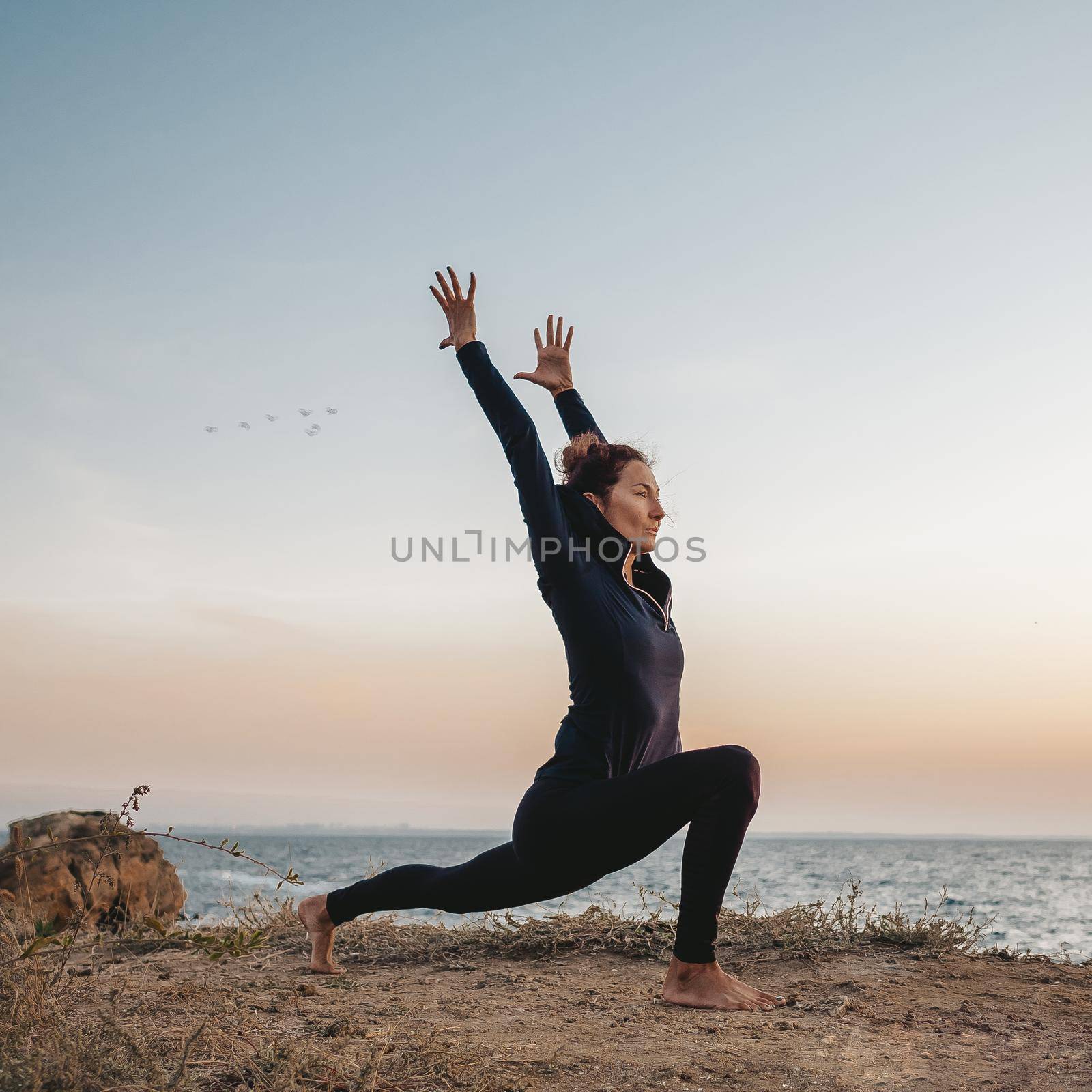 Slim woman in black bodysuit practicing yoga near sea or ocean during sunrise light. Flexibility, stretching, fitness, healthy lifestyle. by kristina_kokhanova
