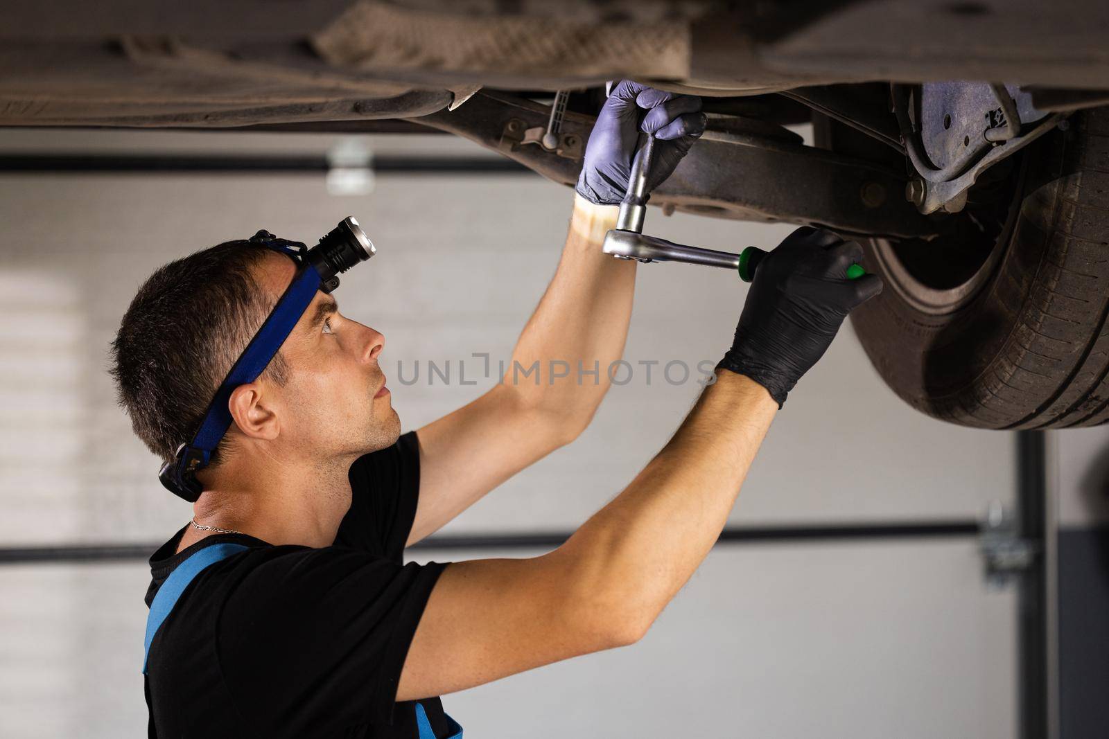 Professional car mechanic screwing details of car with special tool on lifted automobile at repair service station. Skillful man in uniform fixing car. Car service, repair maintenance by uflypro