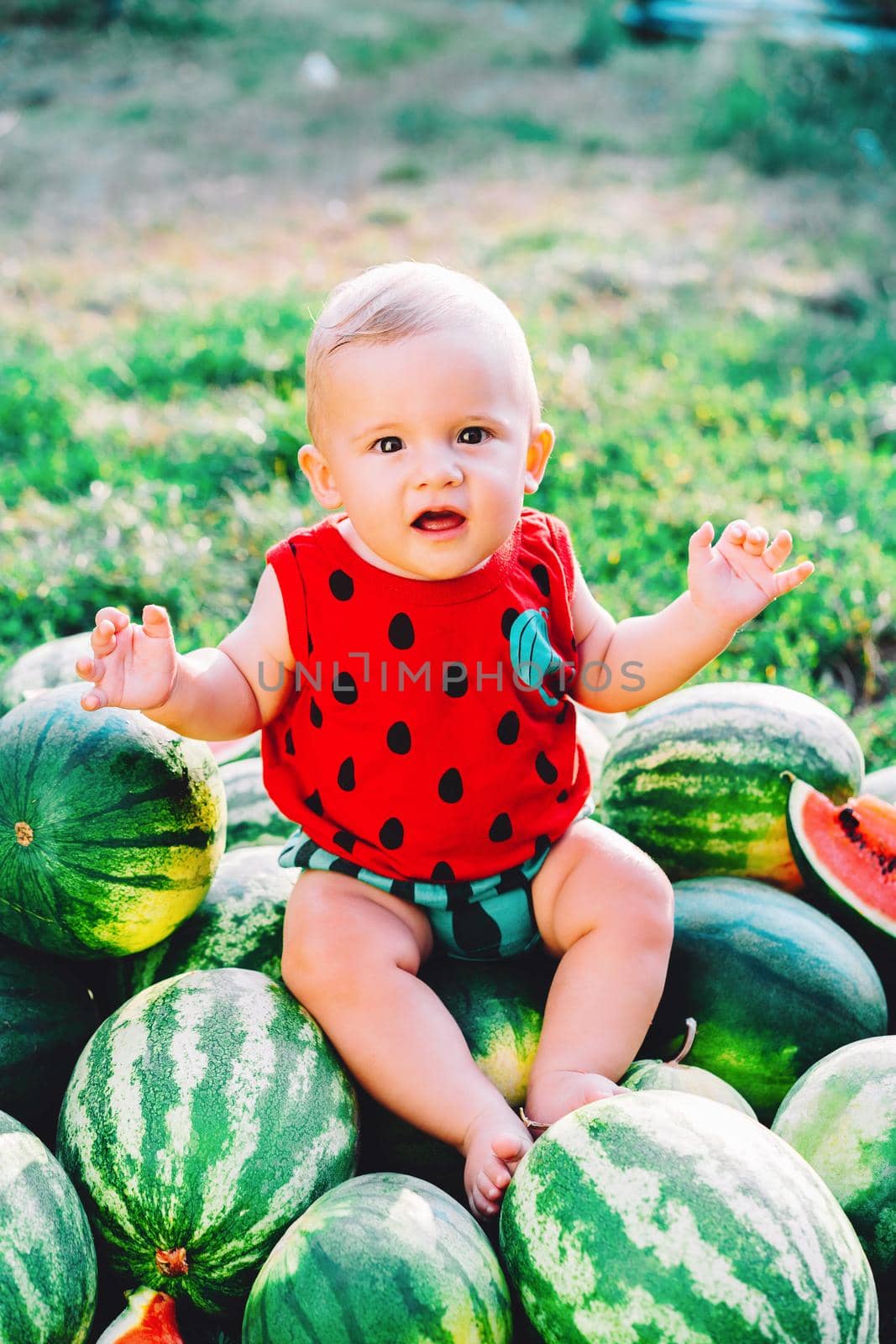 Happy little baby boy sitting on watermelons field or garden. Happy Infant child smiling, looking to camera. Kid on fruits outdoors. Healthy lifestyle concept