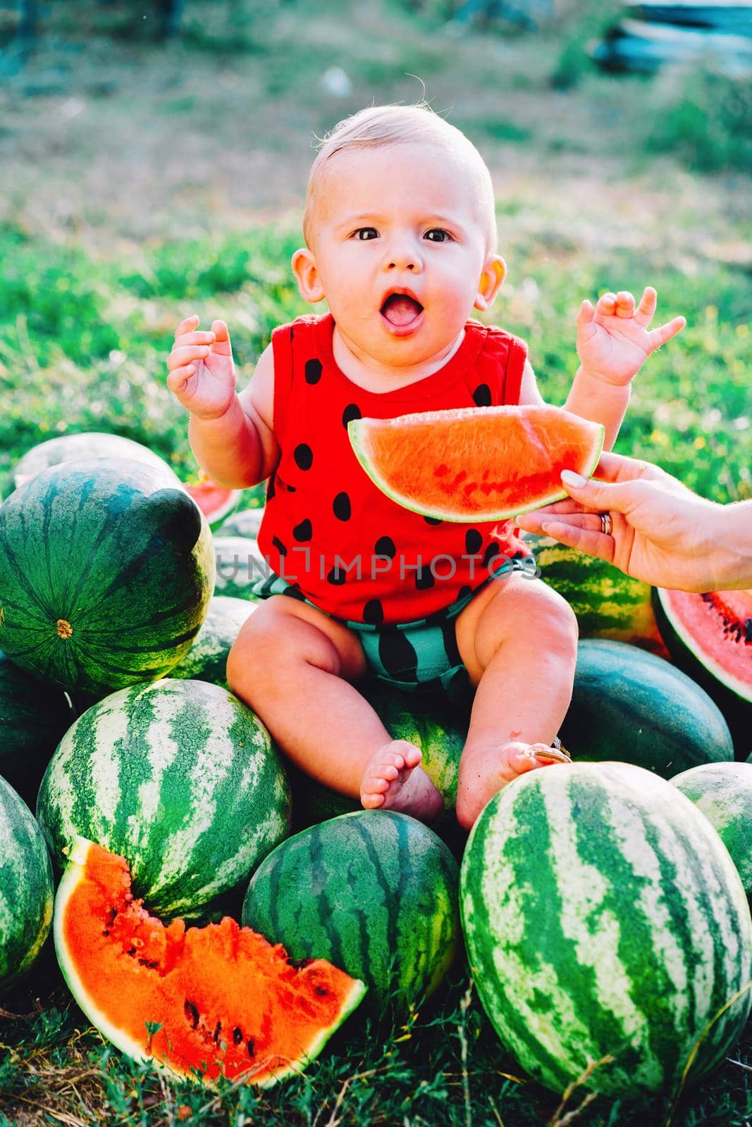 Happy little baby boy in funny costume sitting and eating slice of watermelon on field or garden. Happy Infant child smiling. Kid eat fruit outdoors. by kristina_kokhanova