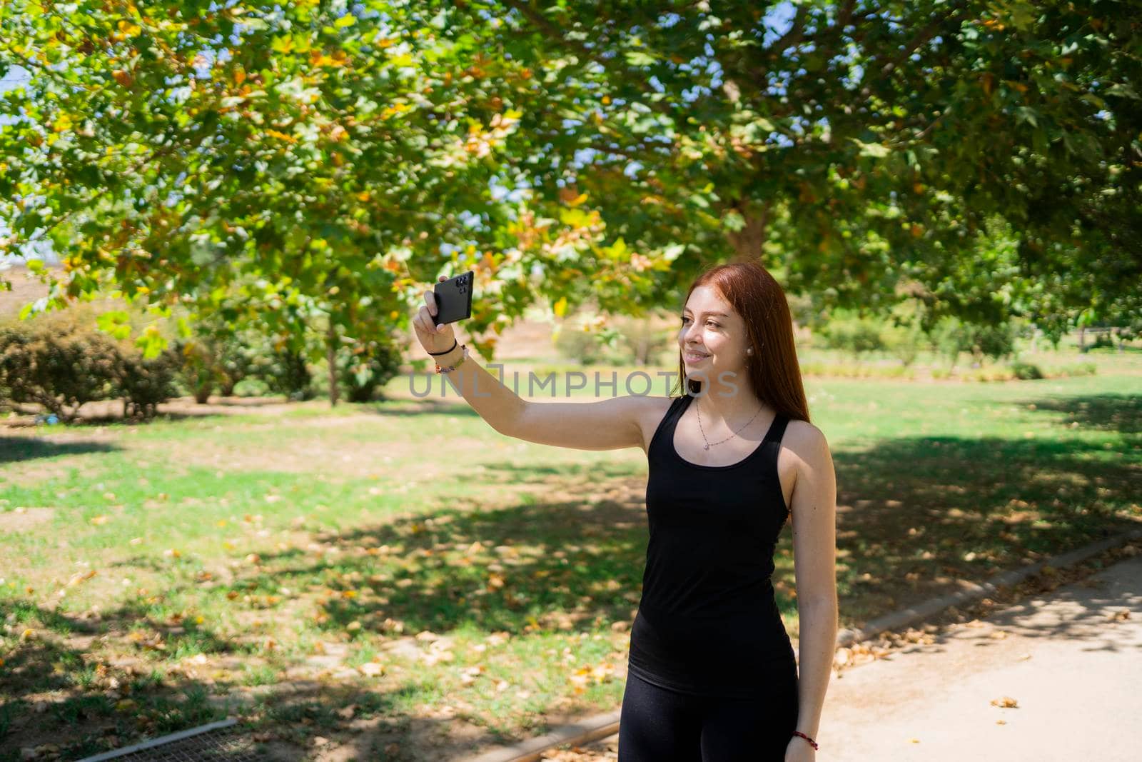 Glad young woman in casual clothes smiling and taking selfie via smartphone while standing on sunlit park alley on summer weekend day