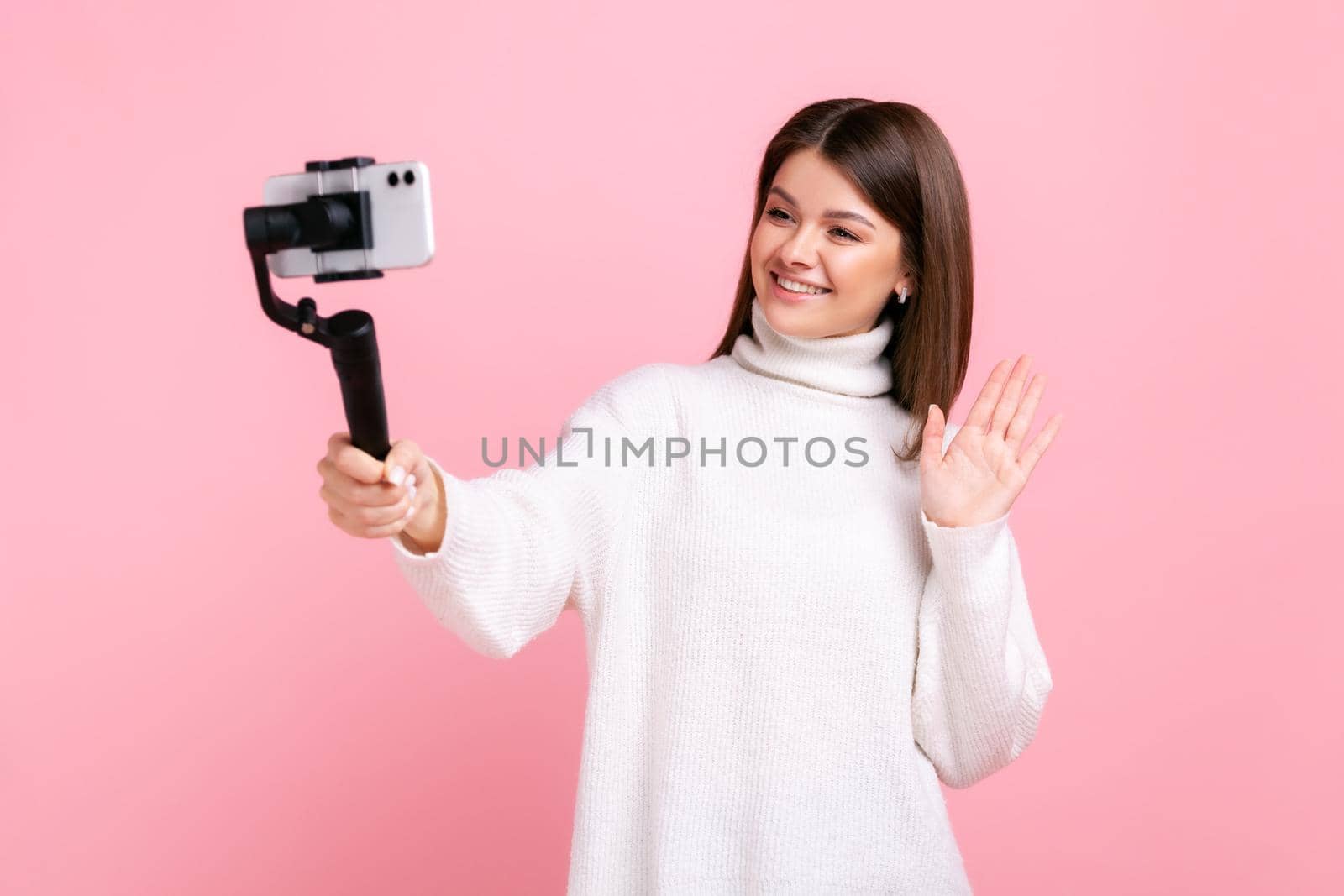 happy woman blogger using smart phone and steadicam for streaming, waving hand, greeting followers, wearing white casual style sweater. Indoor studio shot isolated on pink background.