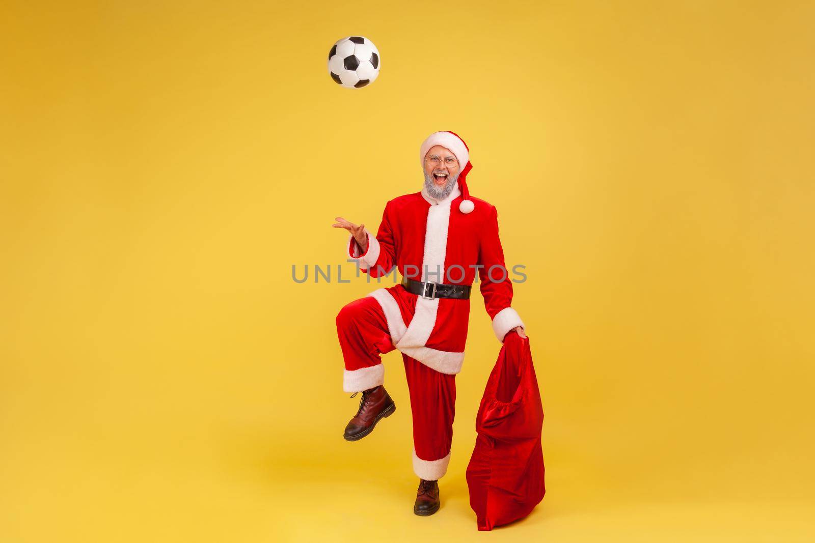 Full length of elderly man with gray beard wearing santa claus costume playing with soccer ball, holding red bag bags with new year presents. by Khosro1