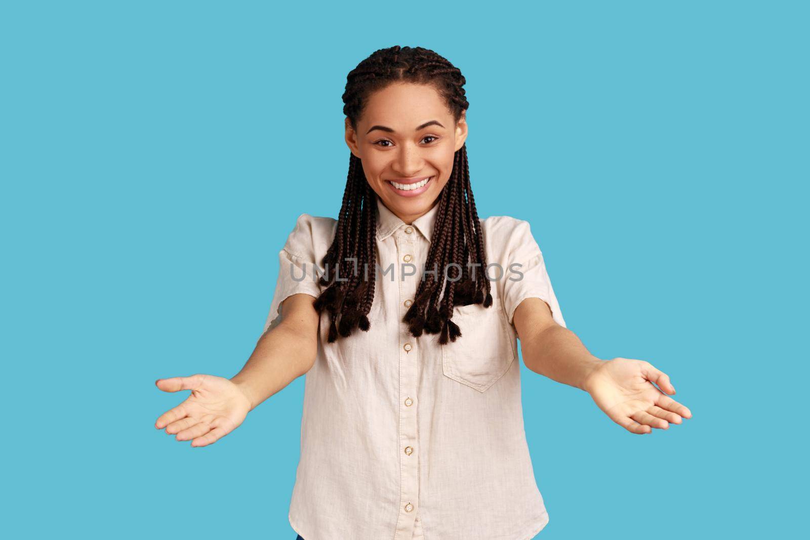 Please, take it. Portrait of kind generous woman with black dreadlocks standing with wide open hands as if sharing for free, giving charity and smiling. Indoor studio shot isolated on blue background.