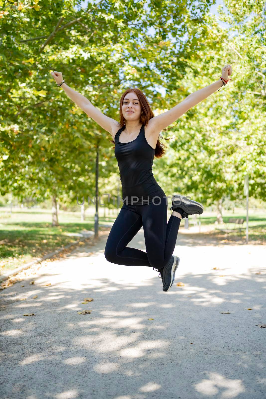 Full body cheerful young female in black clothes raising arms and looking at camera while jumping over asphalt path in sunlit park