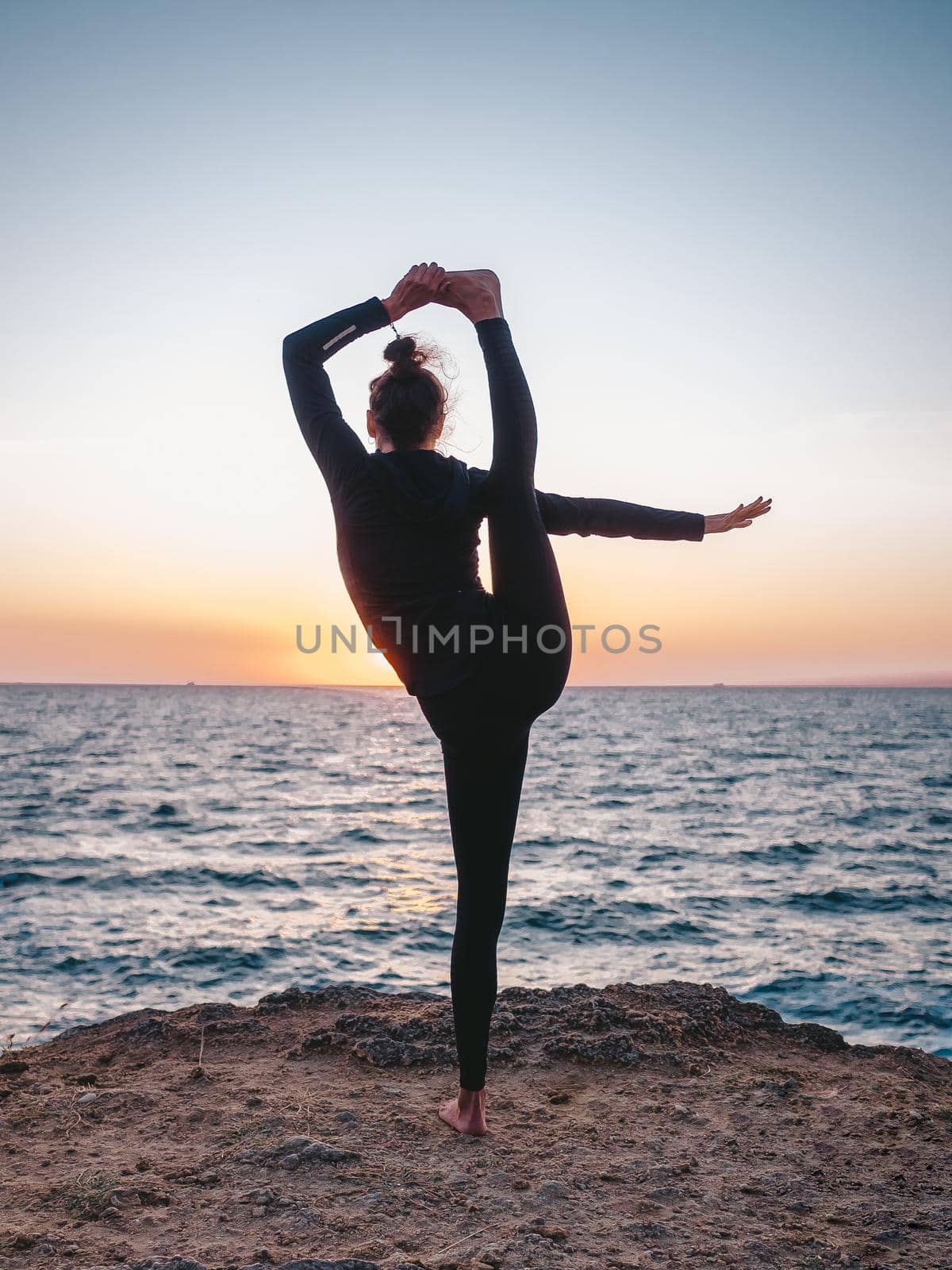 Slim woman in black bodysuit practicing yoga near sea or ocean during sunrise light. Flexibility, stretching, fitness, healthy lifestyle. High quality photo
