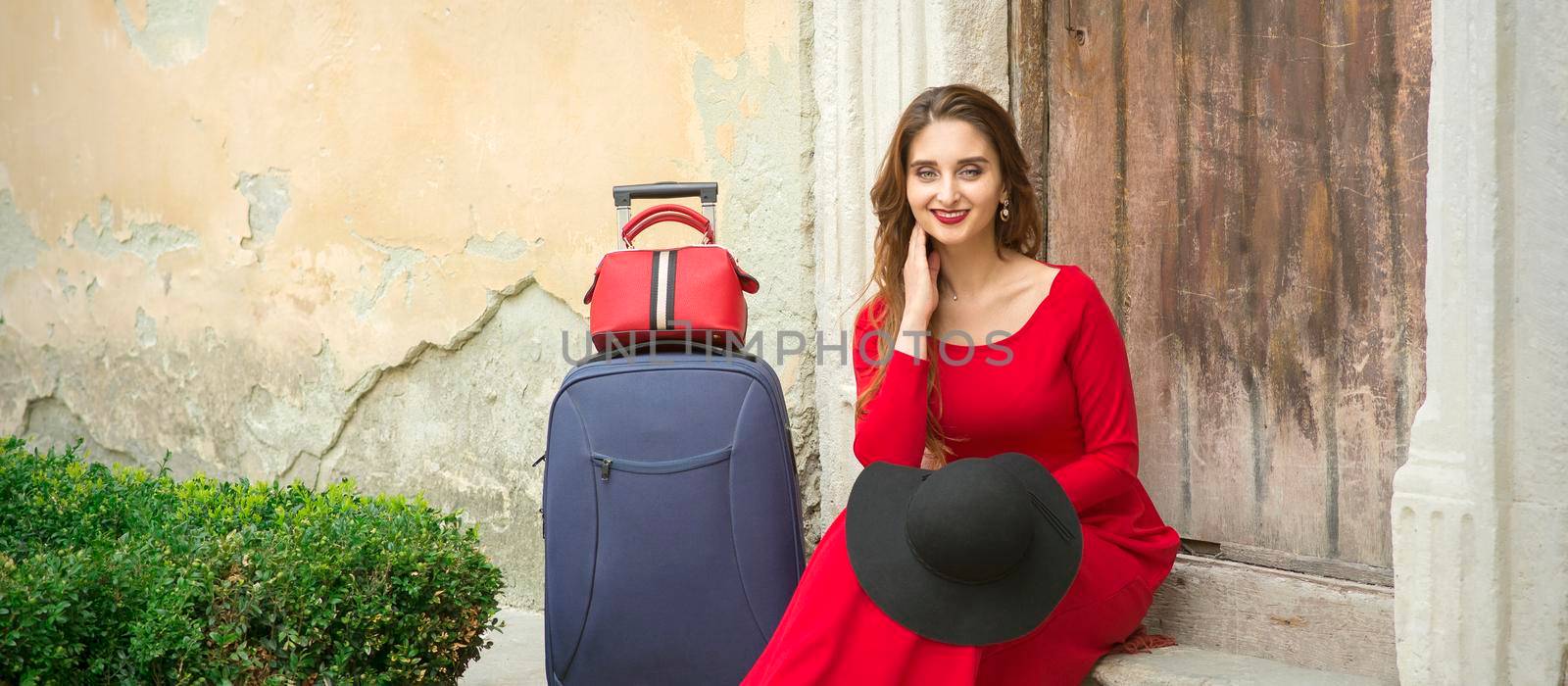 A young caucasian woman is sitting on the threshold of an old house in a red long dress with suitcase looking at the camera outdoors