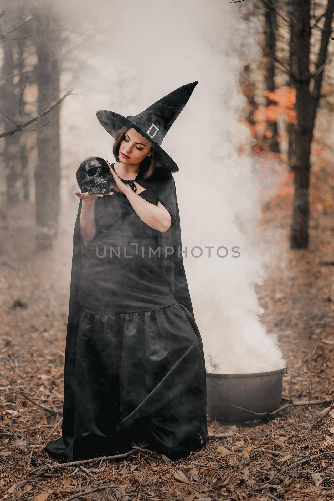 Black witch holding skull in hands, stroking it on autumn forest background. Horror, halloween, cosplay holiday, magic concept. High quality photo