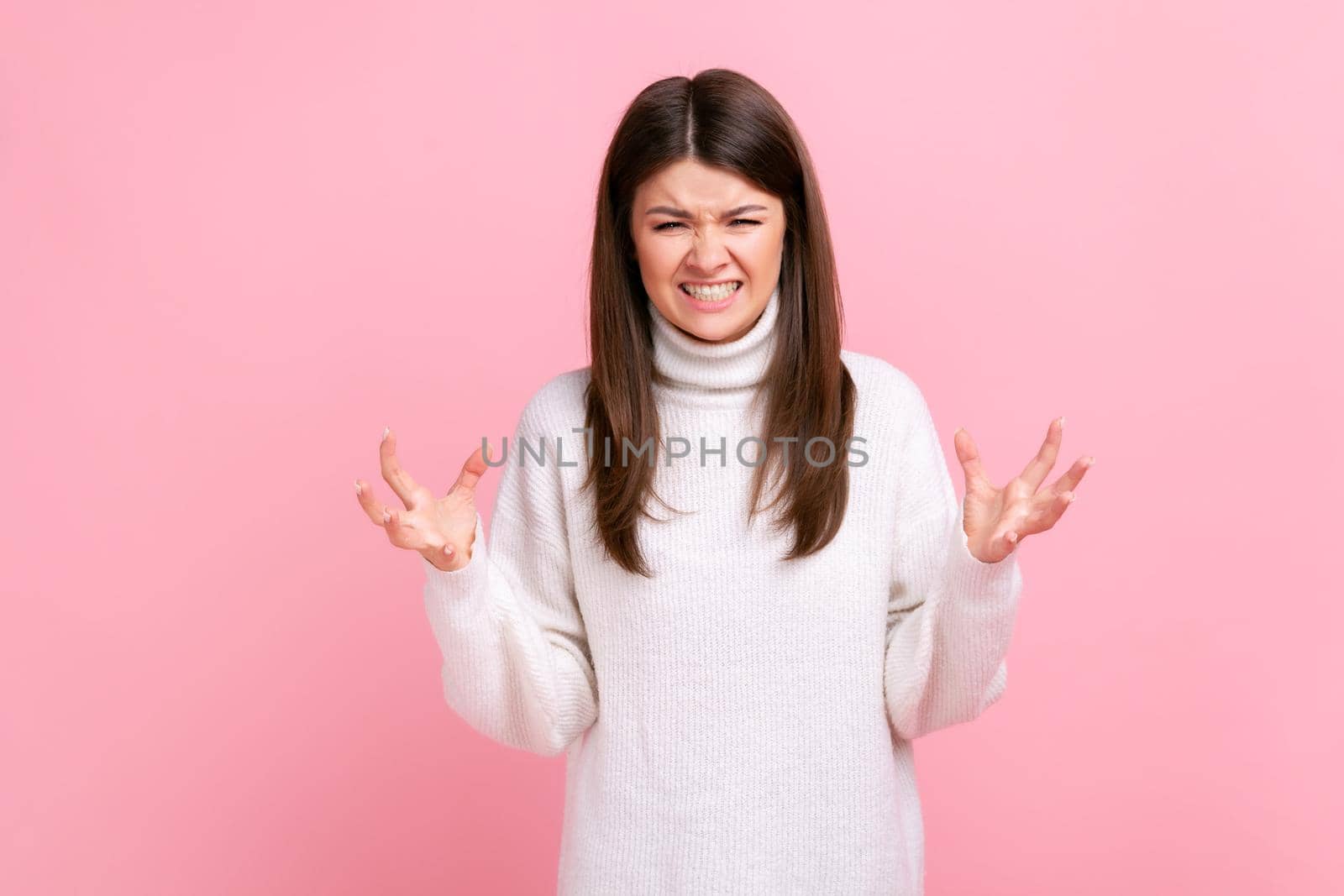 Portrait of angry female standing with raised arms, expressing aggression, screaming, frowning face, wearing white casual style sweater. Indoor studio shot isolated on pink background.