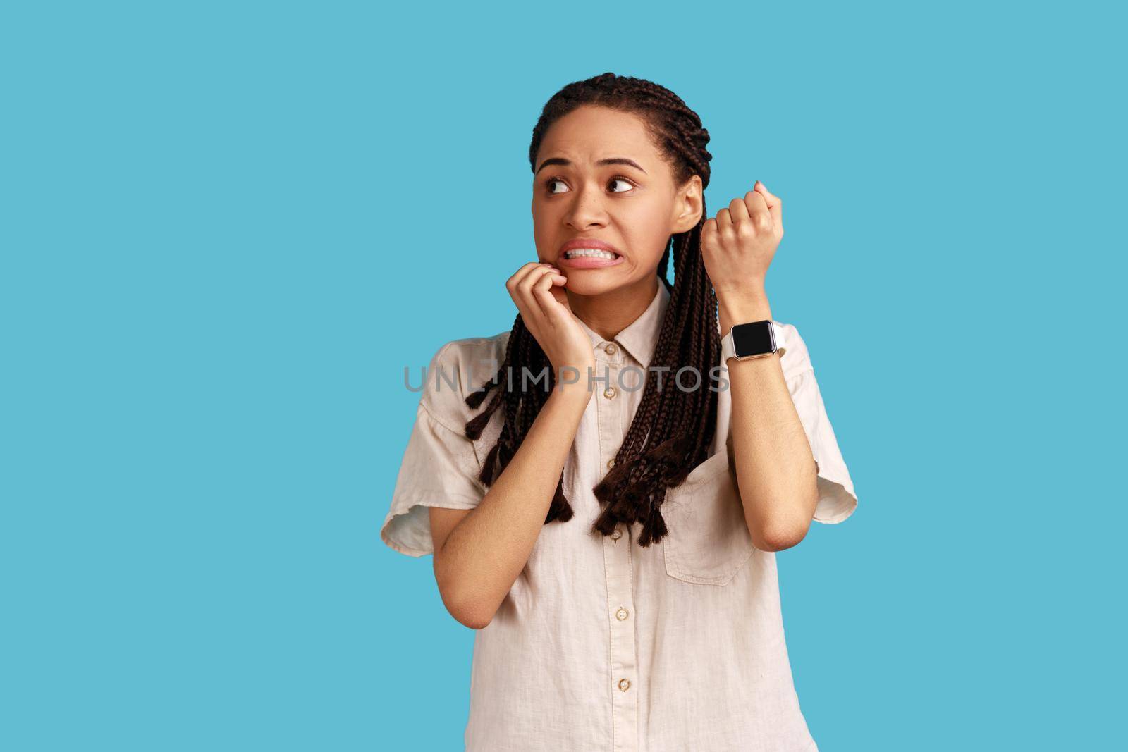 Displeased impatient woman pointing finger on her wrist clock, looking at camera with anxious, worried about deadline, wearing white shirt. Indoor studio shot isolated on blue background.