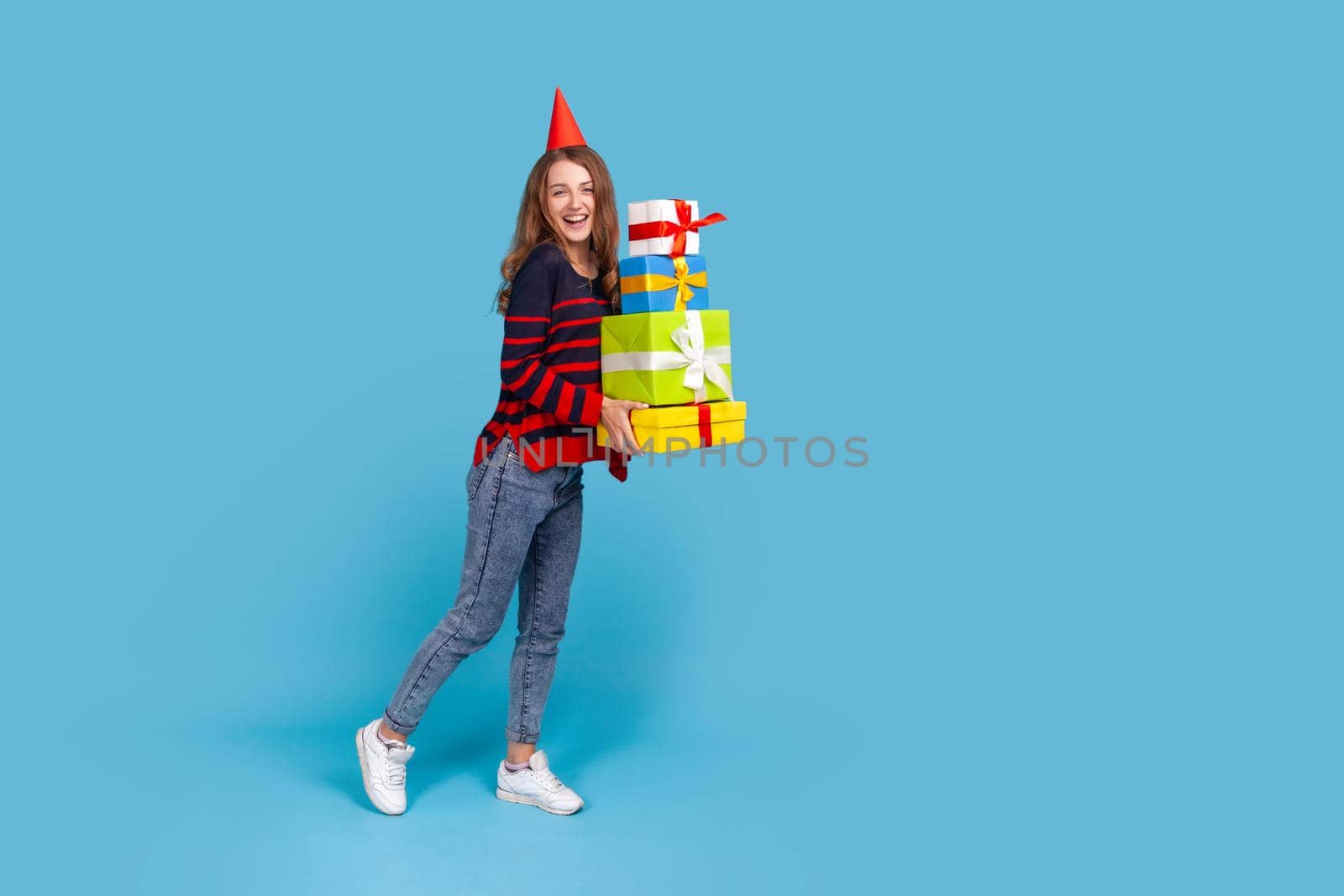 Full length portrait of woman wearing striped casual style sweater and red party box standing with many present boxes, looking at camera with excitement. Indoor studio shot isolated on blue background