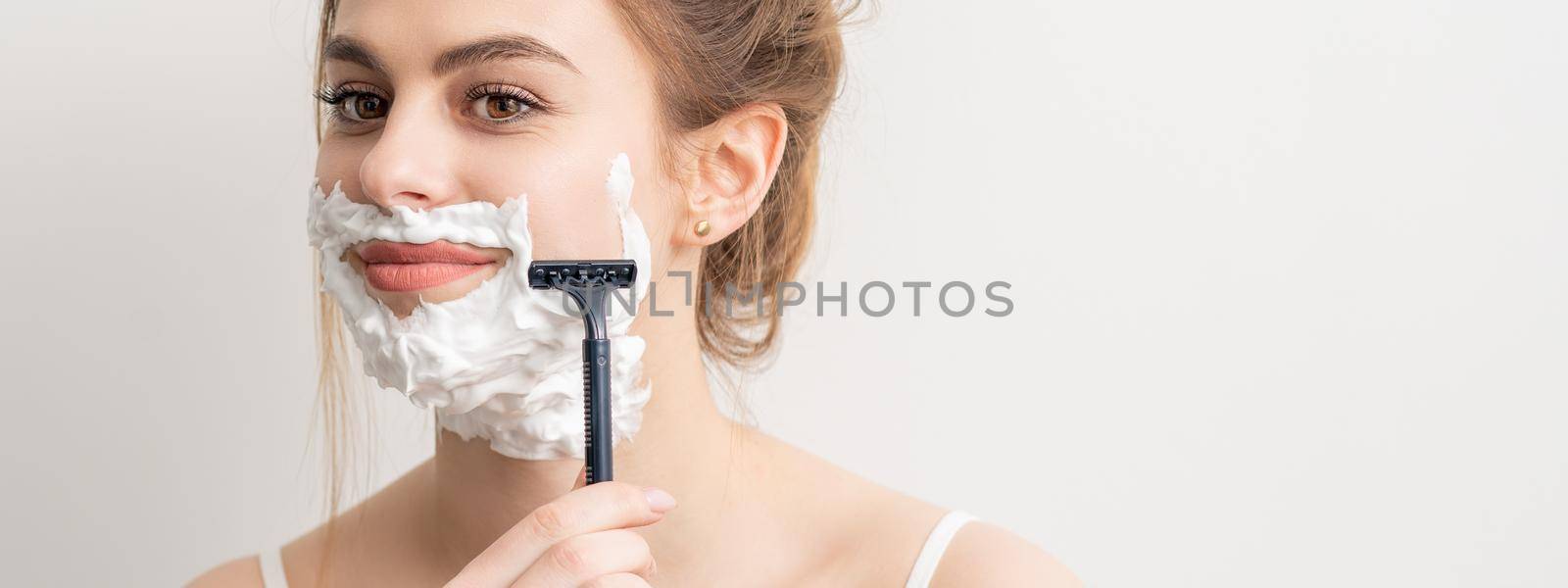 Beautiful young caucasian smiling woman shaving her face with razor on white background