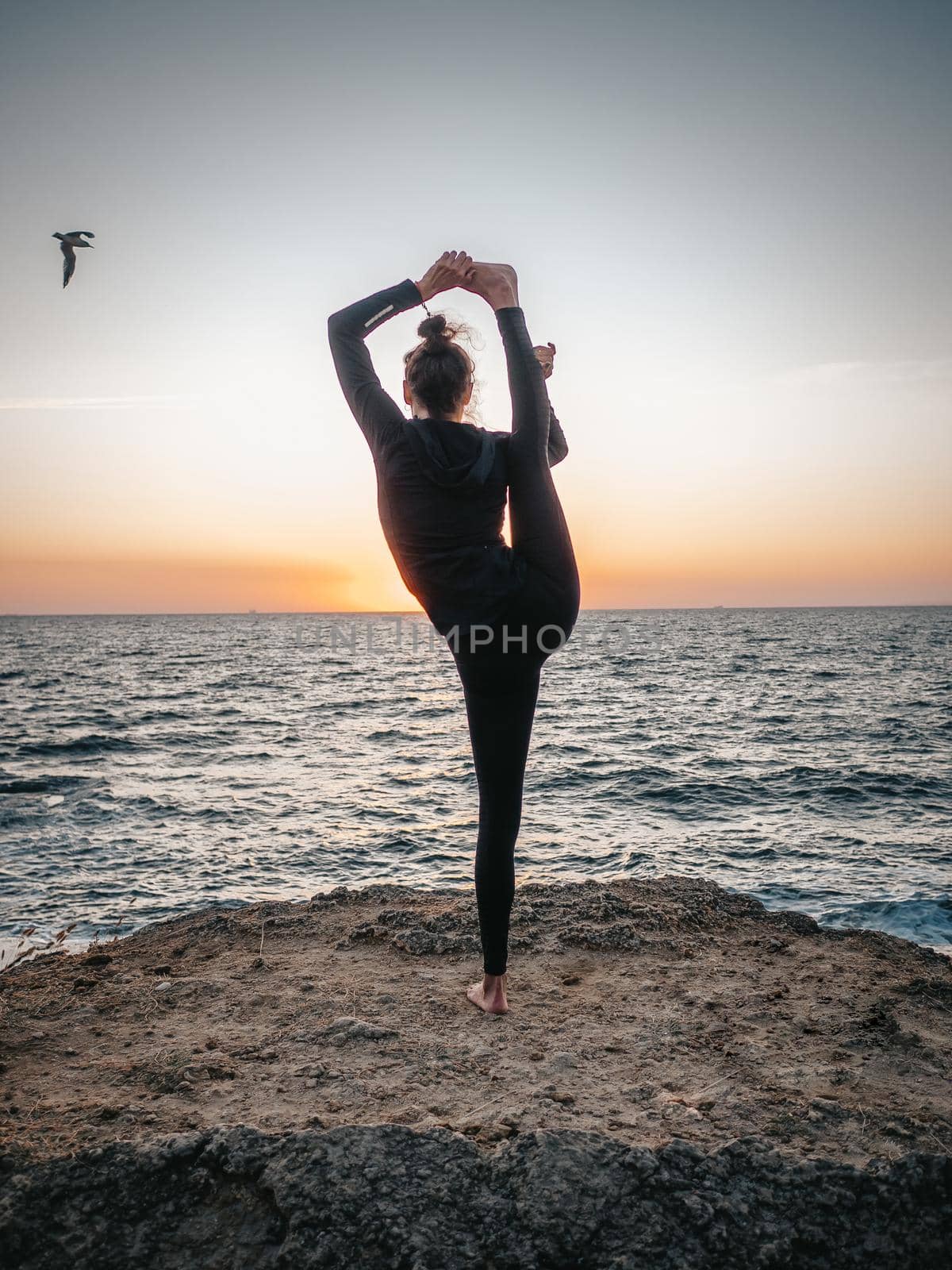 Slim woman in black bodysuit practicing yoga near sea or ocean during sunrise light. Flexibility, stretching, fitness, healthy lifestyle. High quality photo