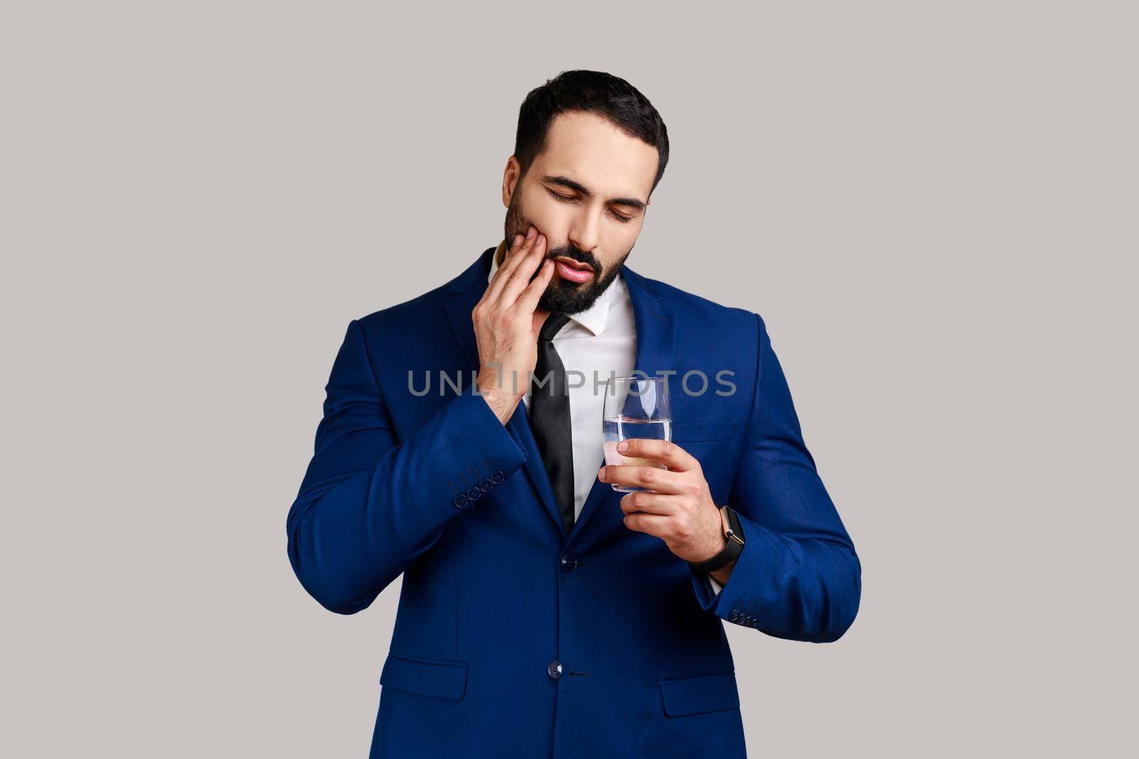 Portrait of sick unhealthy attractive bearded businessman suffering from toothache after drinking cold water, wearing official style suit. Indoor studio shot isolated on gray background.