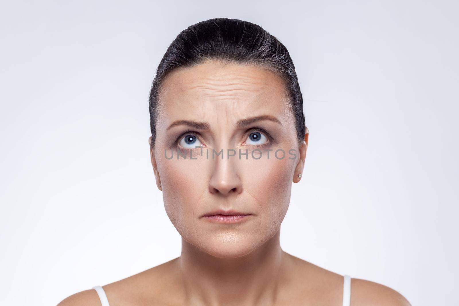 Closeup portrait of well-groomed middle aged woman checking wrinkles on her forehead, looking up, anti aging rejuvenation, skin care. Indoor studio shot isolated on white background.