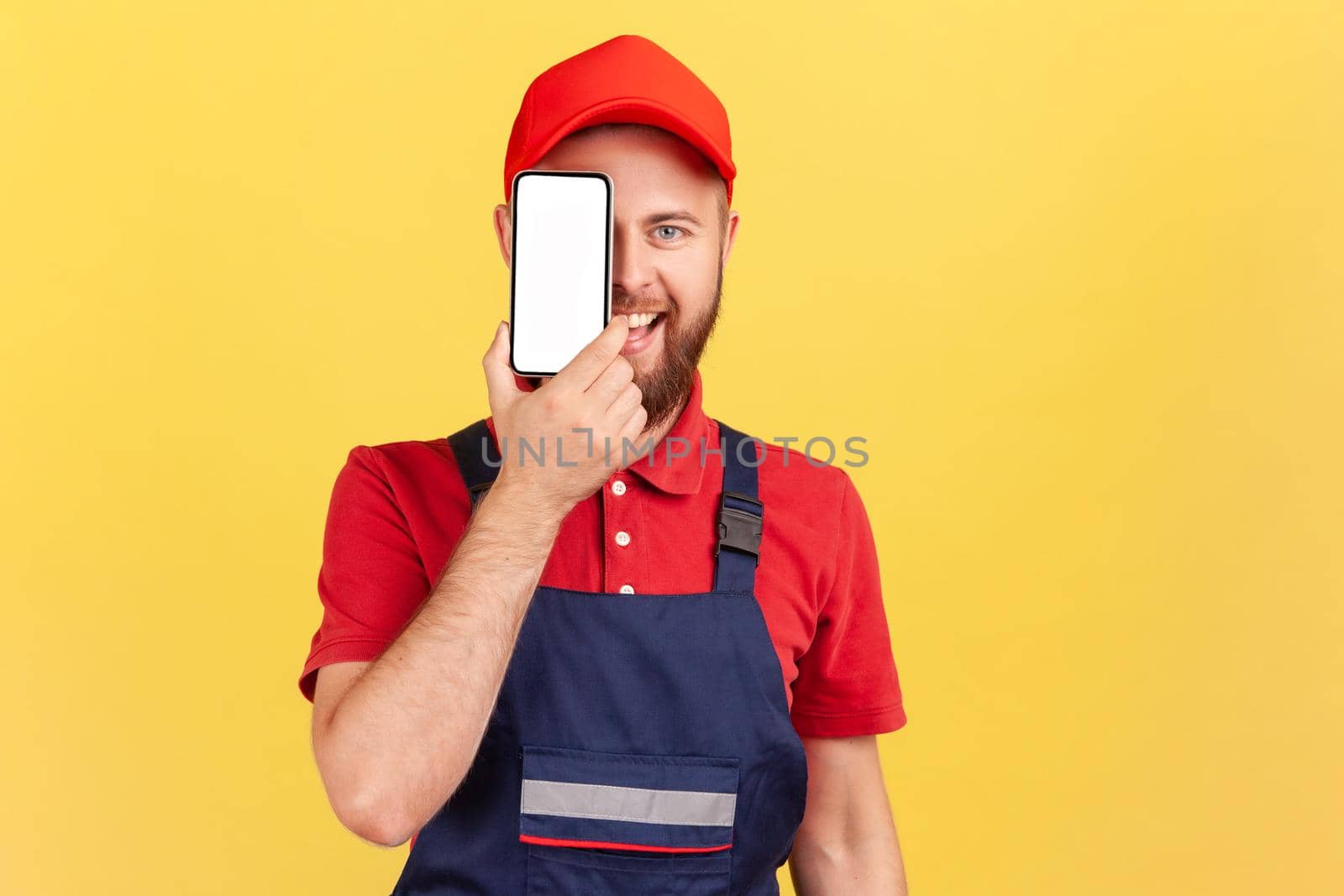 Portrait of happy smiling joyful bearded worker man standing and covering eye with cell phone with blank screen for promotional text. Indoor studio shot isolated on yellow background.