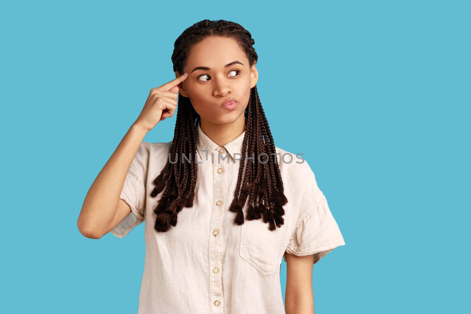 Positive woman with black dreadlocks holding finger near head temple and gesturing stupid idiot, looking displeased with crazy idea, wearing white shirt. Indoor studio shot isolated on blue background