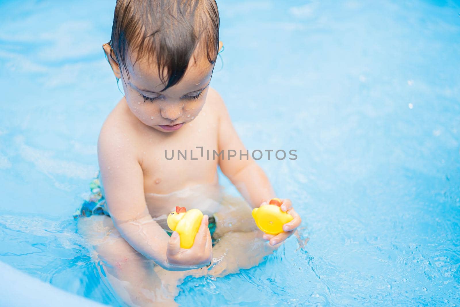 Cute little child bathing with duck in blue street pool in courtyard. Portrait of joyful toddler, baby. Kid laughs, splashes water, smiles. Concept of healthy lifestyle, family, leisure in summer by kristina_kokhanova