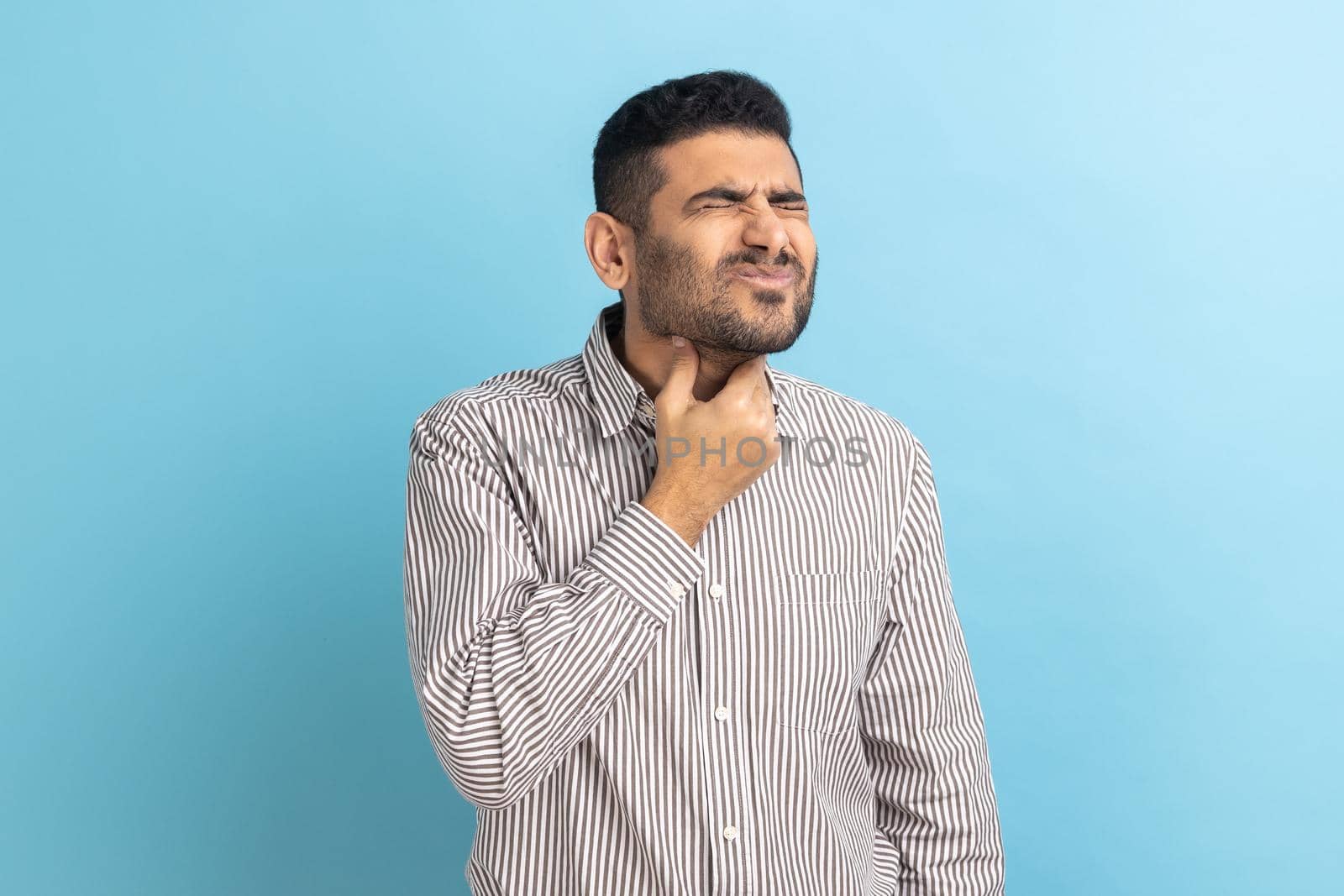 Throat pain. Portrait of unhappy ill businessman touching her neck, suffering sore throat, viral infection or flu symptoms, wearing striped shirt. Indoor studio shot isolated on blue background.