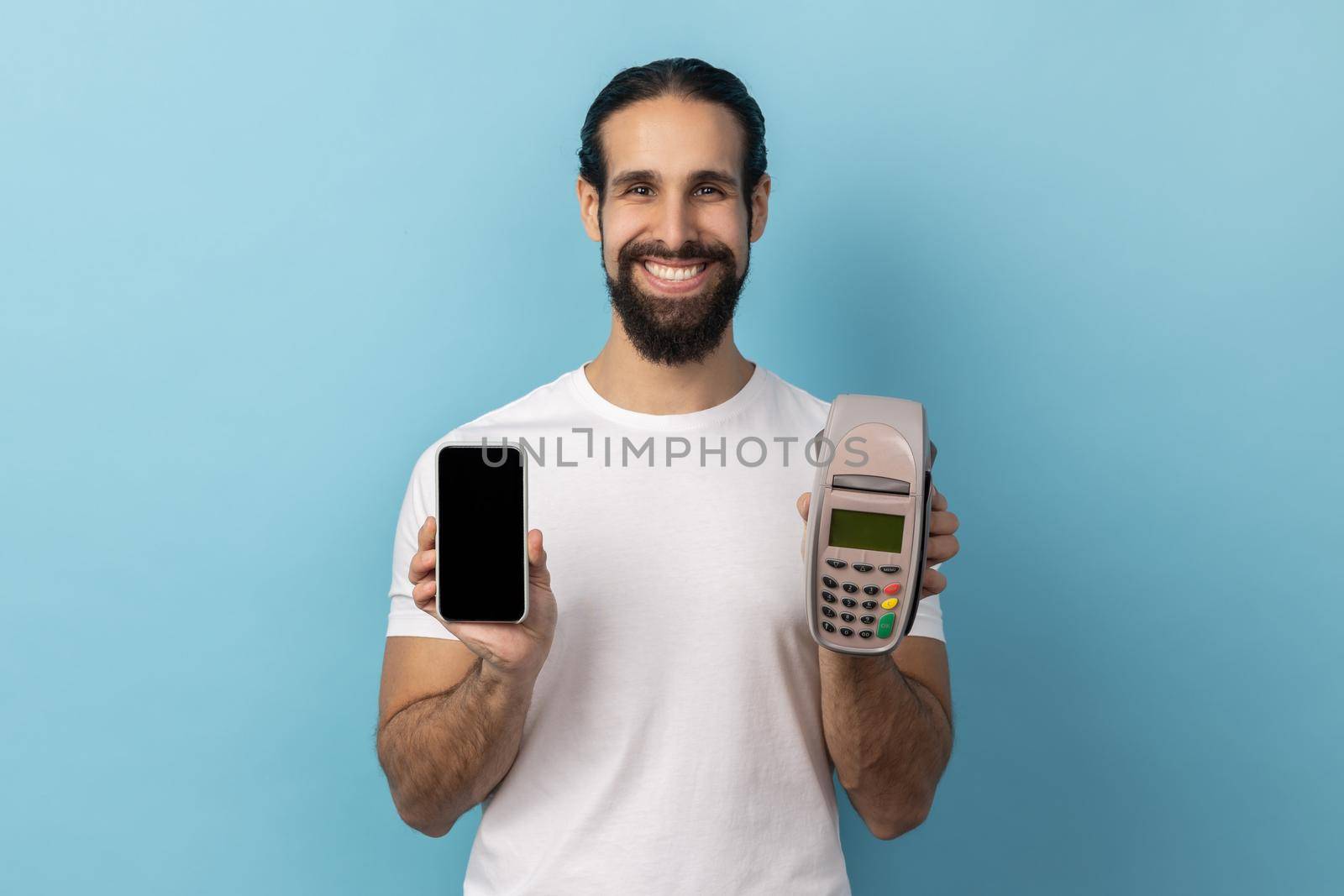 Portrait of smiling happy man with beard wearing white T-shirt holding pos terminal and smart phone with empty display for advertisement. Indoor studio shot isolated on blue background.