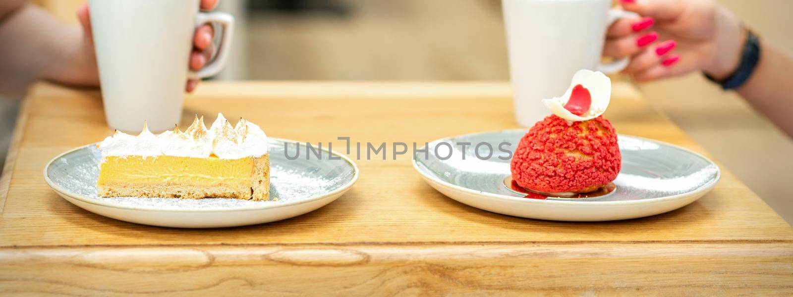 Two young girlfriends drinking coffee with pieces of cake sitting at the table in a cafe outdoors