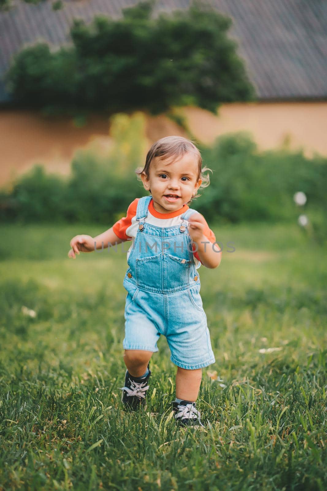 Cheerful perky toddler boy runs to meeting to camera with smile and happiness. Baby boy in denim overall in green park or garden. High quality photo