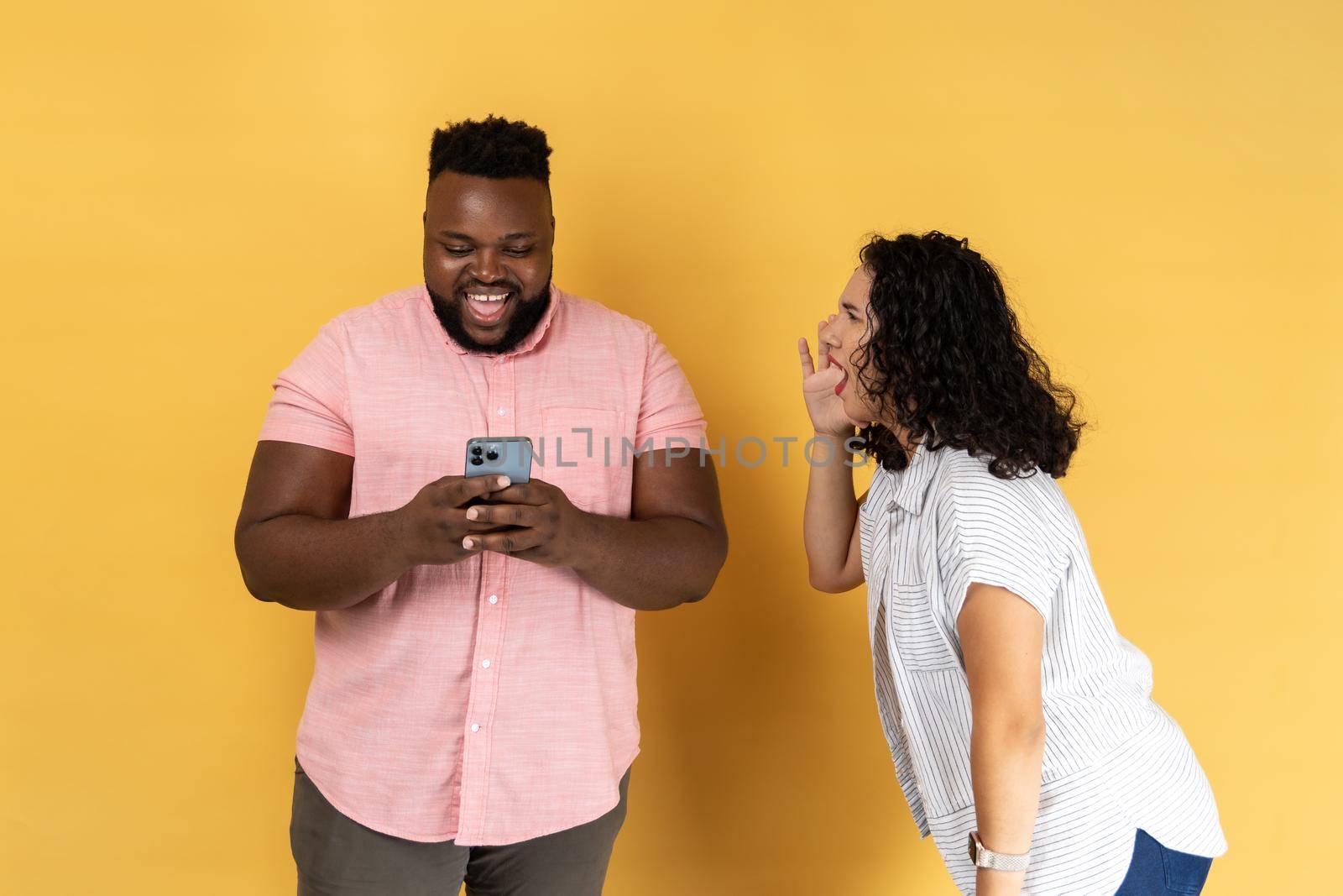 Young couple in casual clothing standing together, woman desperately screaming near happy man using phone and ignoring her, attracting his attention. Indoor studio shot isolated on yellow background.