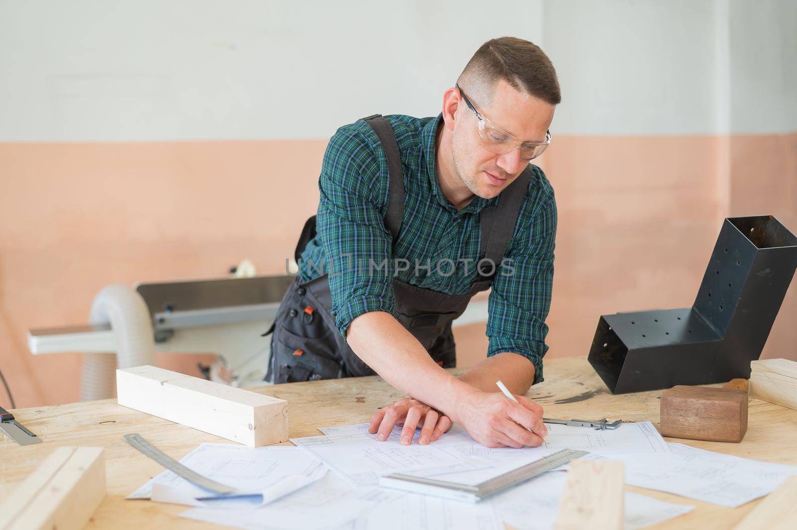 A portrait of a carpenter in goggles and work overalls draws a blueprint for a workshop