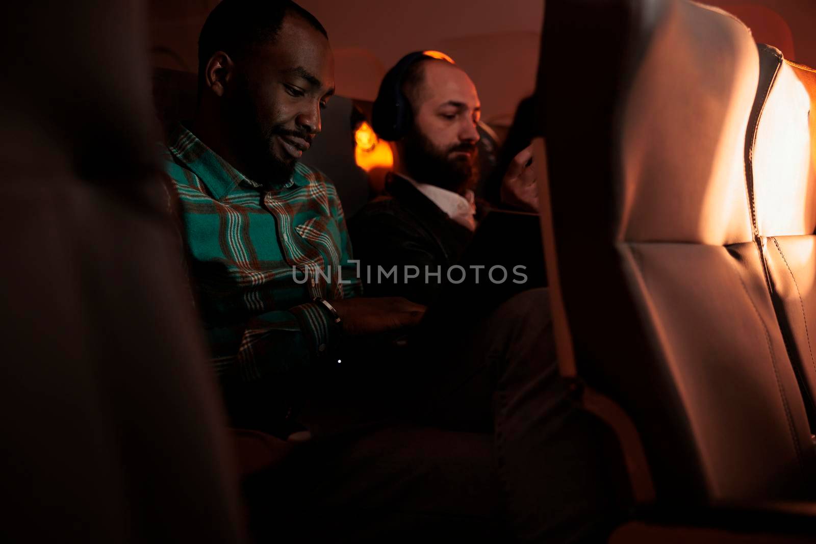 Group of diverse people travelling to destination by airplane, using computer or mobile phone on flight during sunset. Flying in economy class with tourists to go on holiday vacation or work trip.