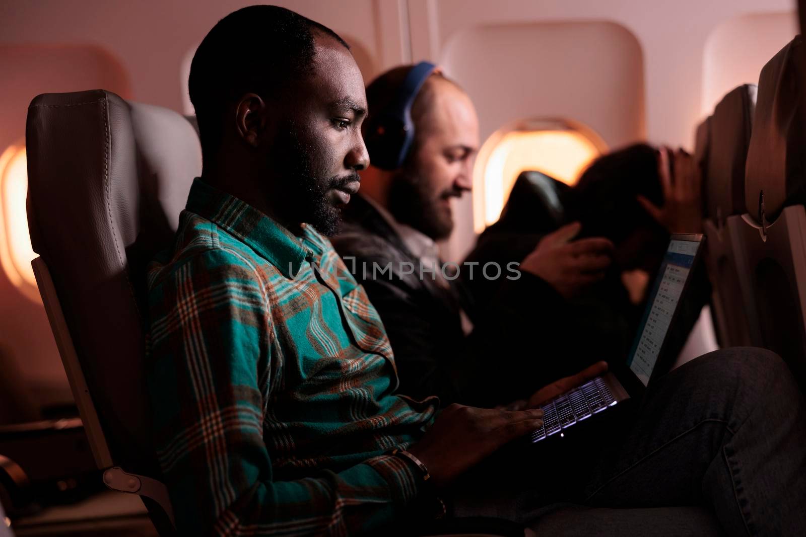 Male employee travelling in economy class and using laptop during flight, flying abroad on holiday destination or work trip. Working on computer during sunset, aerial transportation.