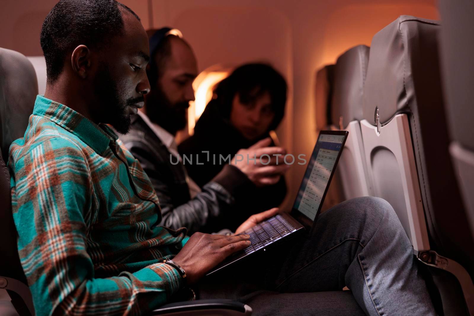 Male passenger travelling abroad by airplane on international flight, using airline service and aerial transportation. Freelancer using laptop computer during sunset in economy class.