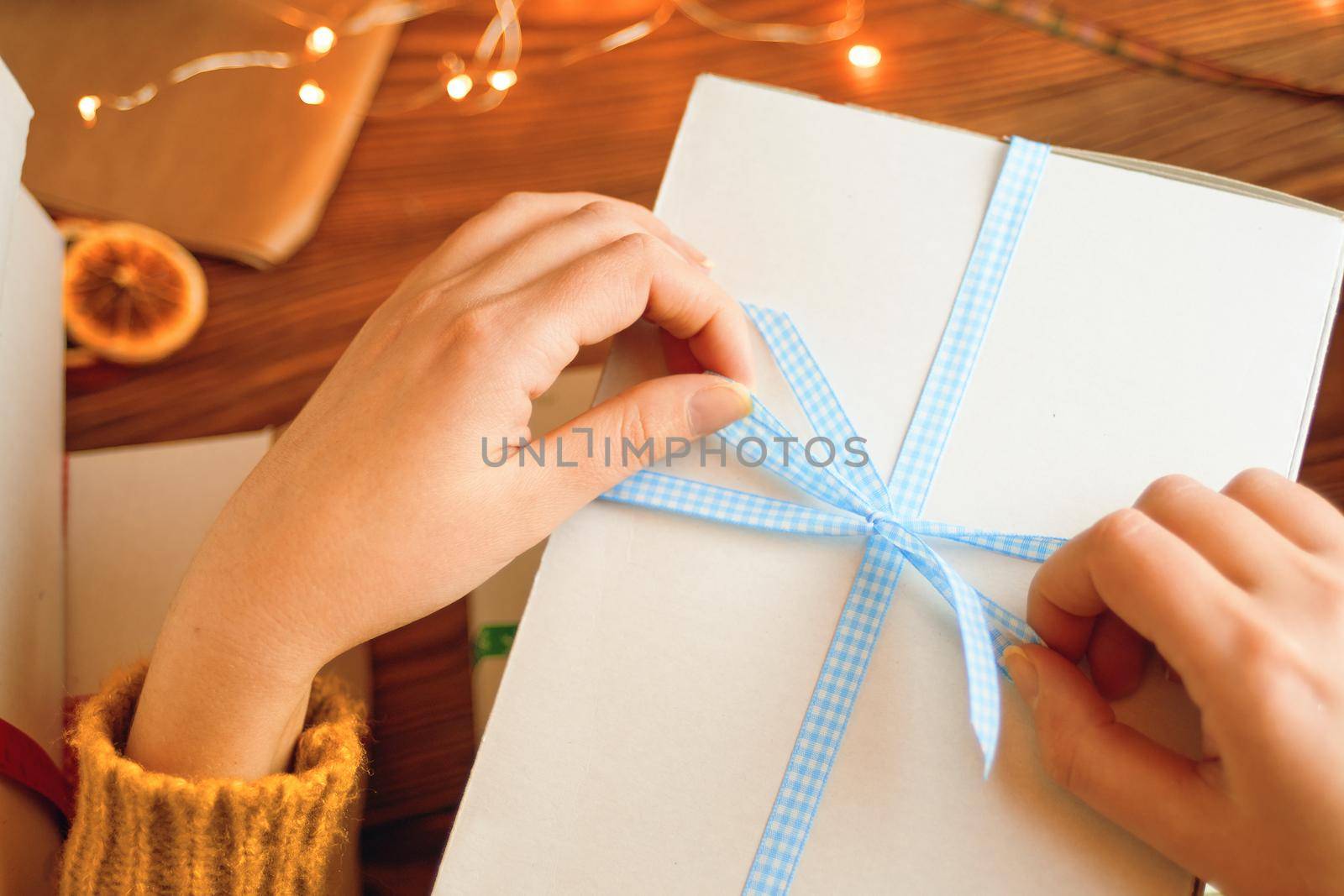 Girl's hands in orange knitted sweater opening white gift box. New Year's festive atmosphere. Christmas surprise with ribbon and garland lights on wooden table.