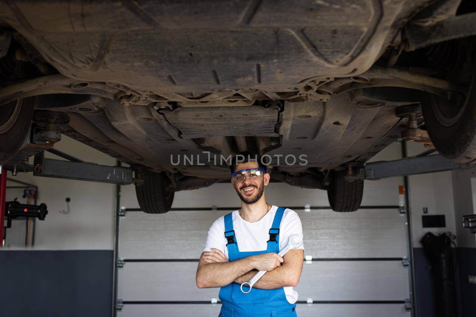 Mechanic man standing in front of the garage. Joyful young bearded man motor mechanic in overalls with tool standing in workshop. Motor mechanic. Concept of small business, own business by uflypro