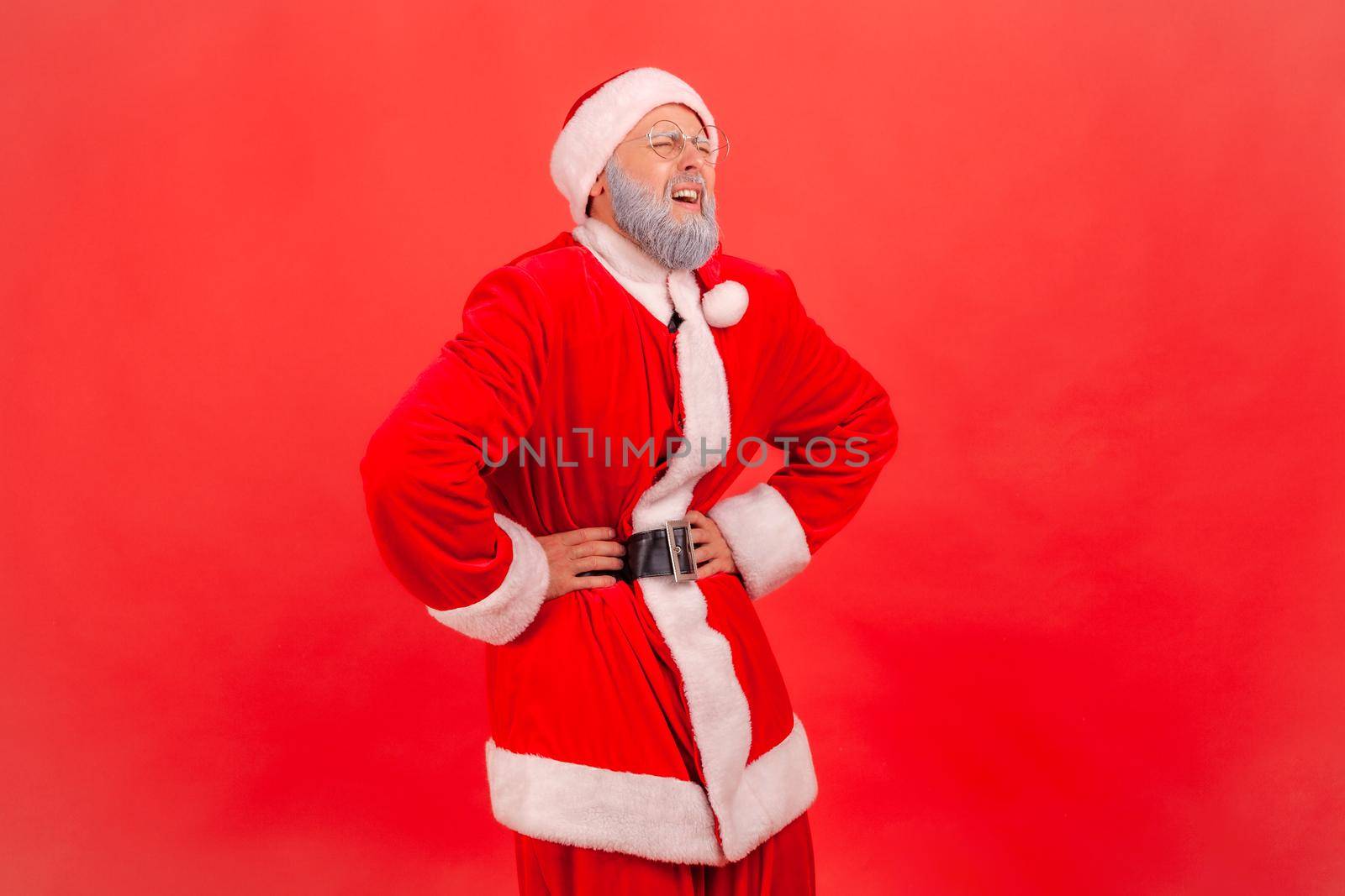 Portrait of elderly man with gray beard wearing santa claus costume standing and keeping hands on belly, suffering from pain, gastritis or constipation. Indoor studio shot isolated on red background.