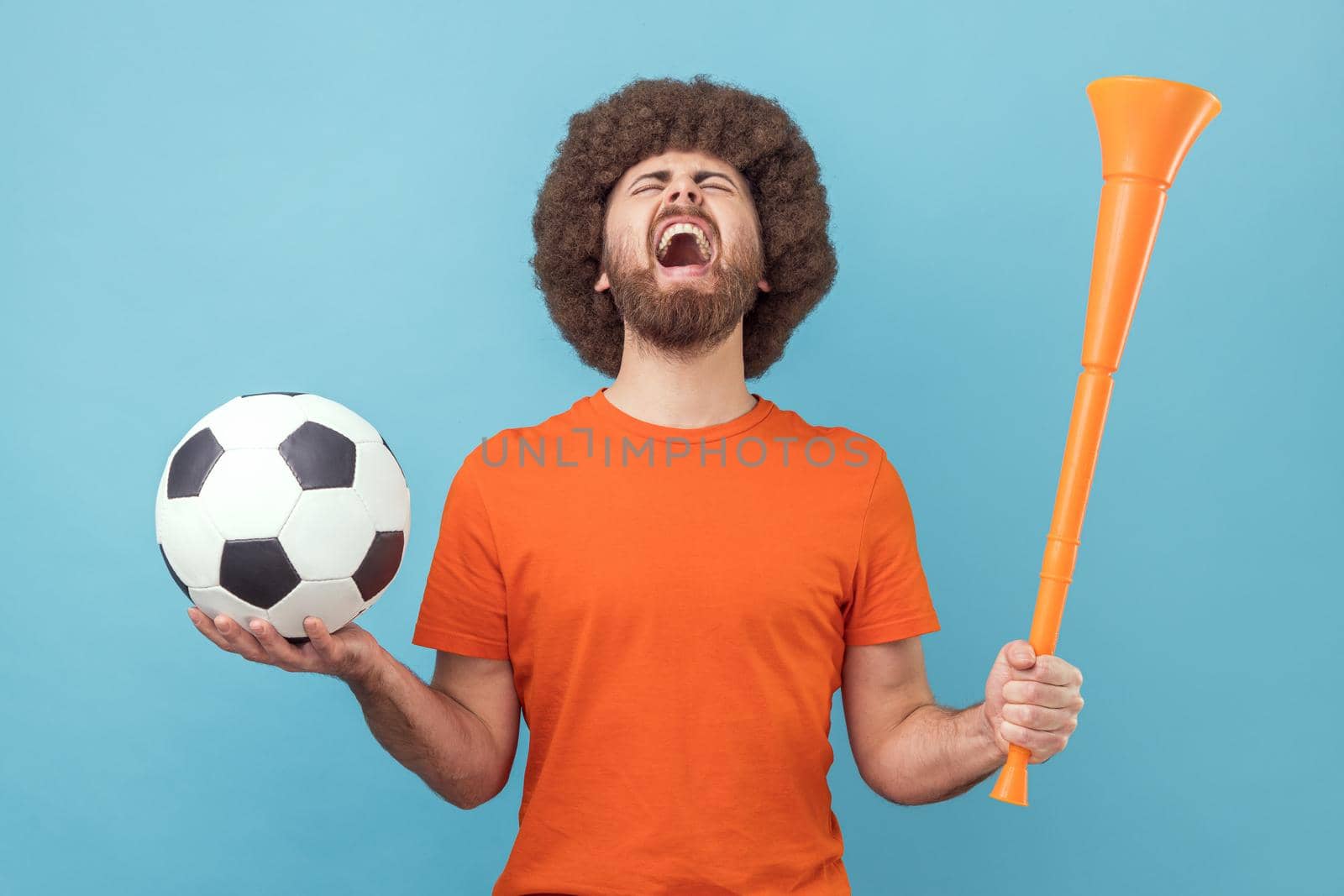 Man with Afro hairstyle wearing T-shirt screaming widely opening mouth, celebrating victory of favourite football team, holding horn and ball in hands. Indoor studio shot isolated on blue background.