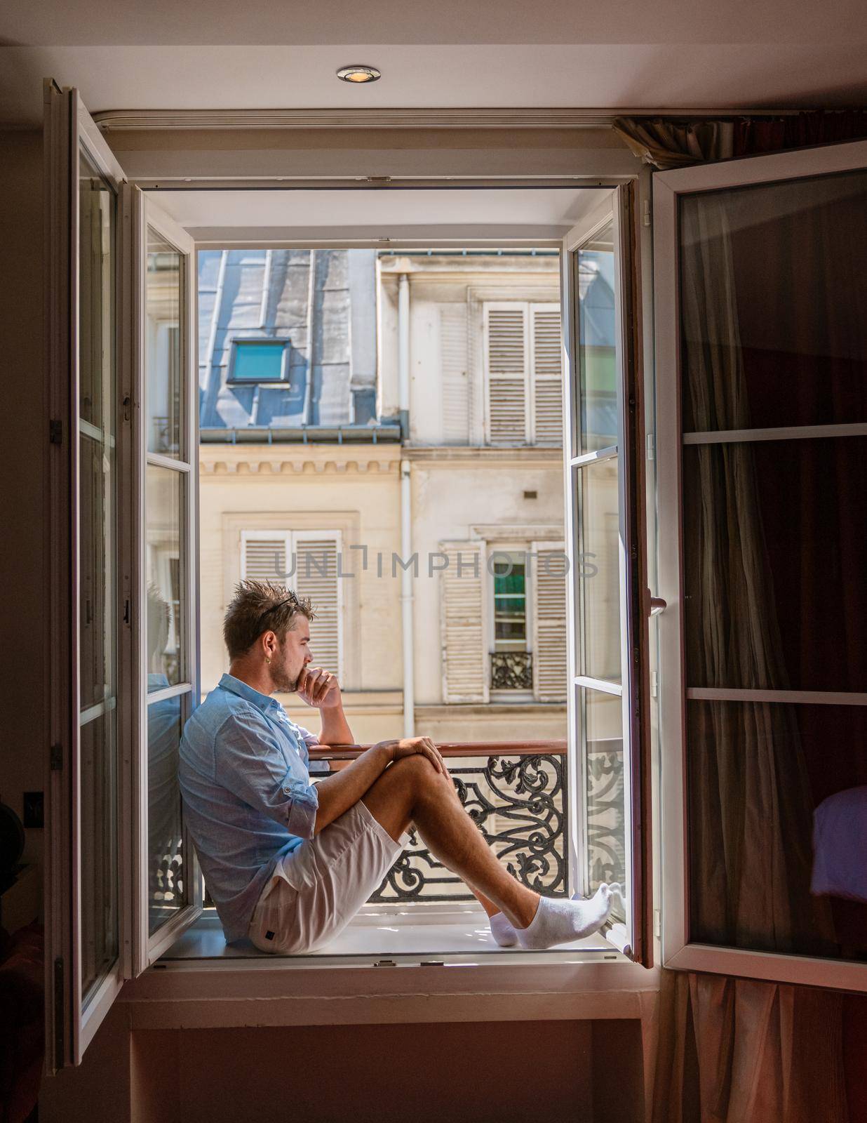 young man sitting in a window looking out over the city Paris. Men enjoy sun in window of hotel room by fokkebok
