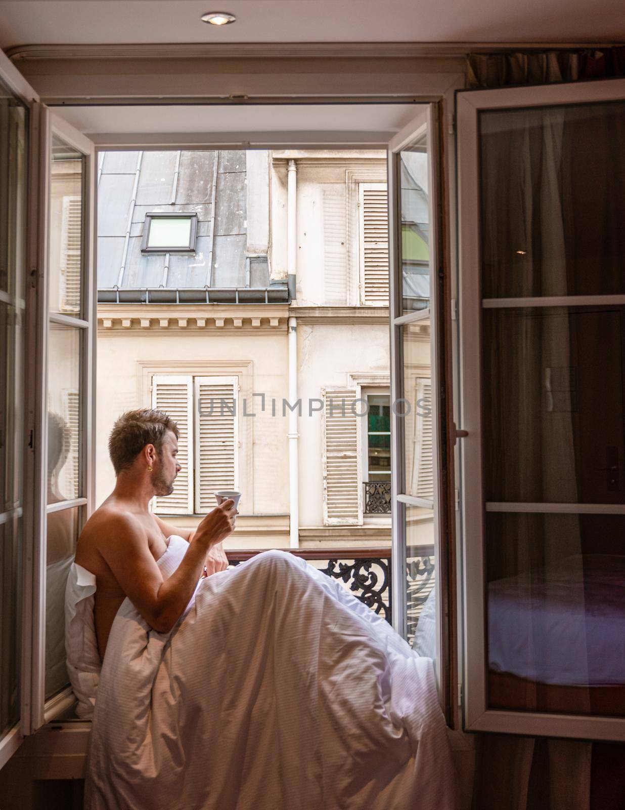 young man sitting in a window looking out over the city Paris. Men enjoy sun in window of hotel room by fokkebok