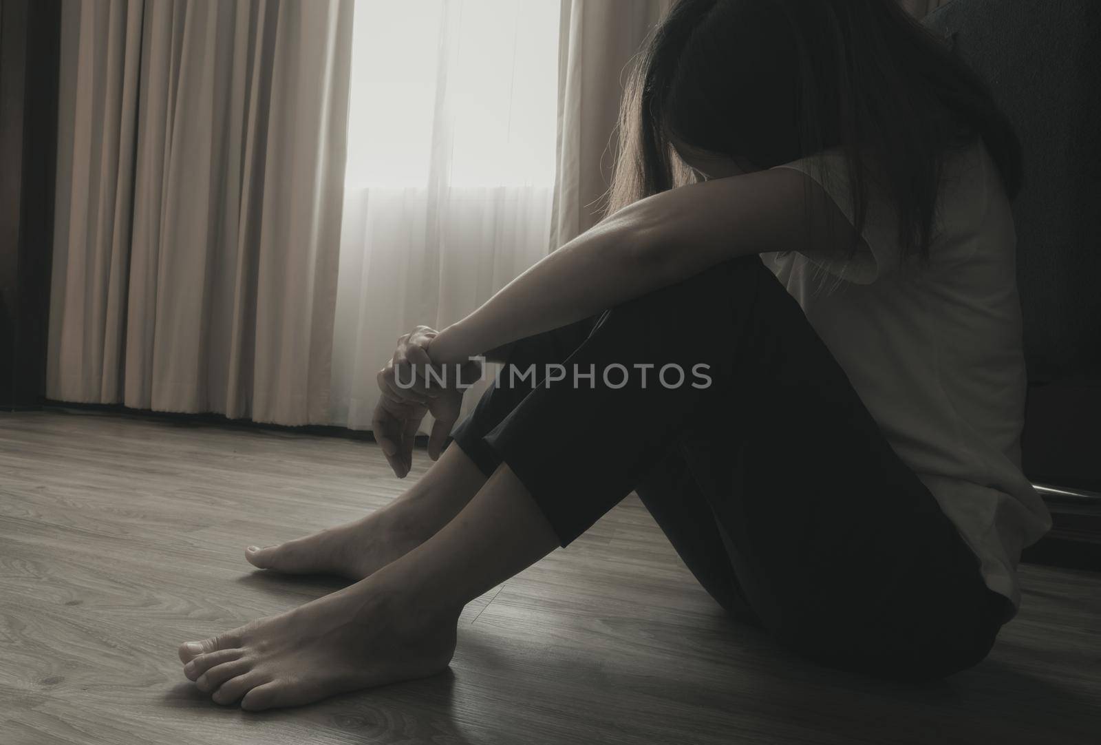 Depressed and stressed woman sitting on the floor near curtains in bedroom. Sad woman sitting in the dark room. Girl with mental health problems. Unhappy life. World Mental Health Day concept.