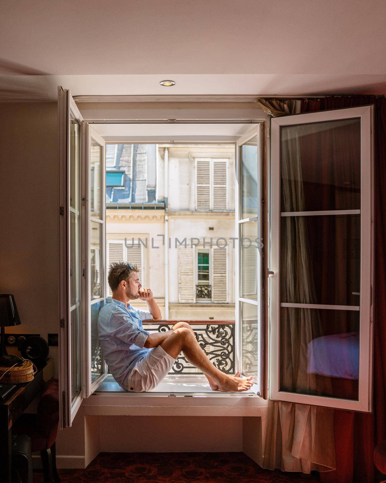 young man sitting in a window looking out over the city Paris. Men enjoy sun in window of hotel room by fokkebok