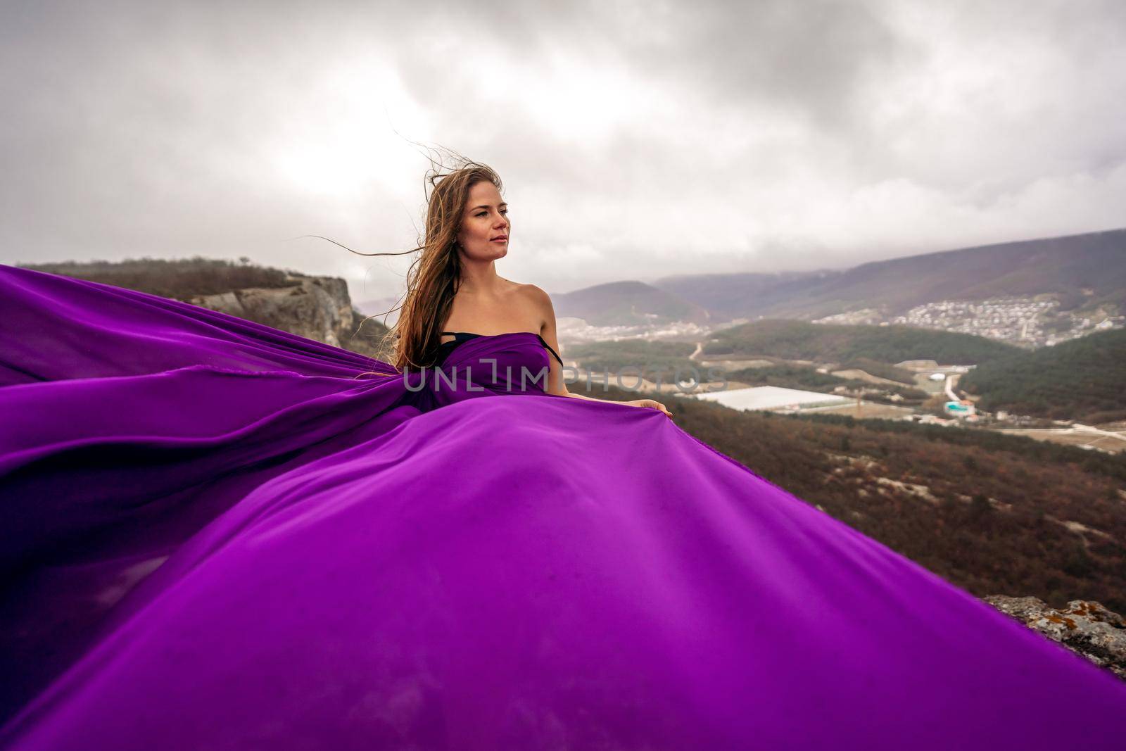 A woman with long hair is standing in a purple flowing dress with a flowing fabric. On the mountain against the background of the sky with clouds. by Matiunina