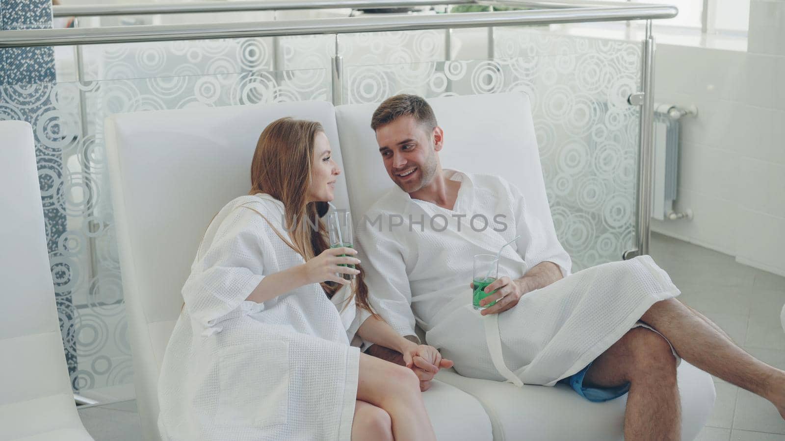 Young lovers in bathrobes are relaxing in chairs in spa salon, drinking cocktails and talking. Wellness, romantic relationship and happiness concept.