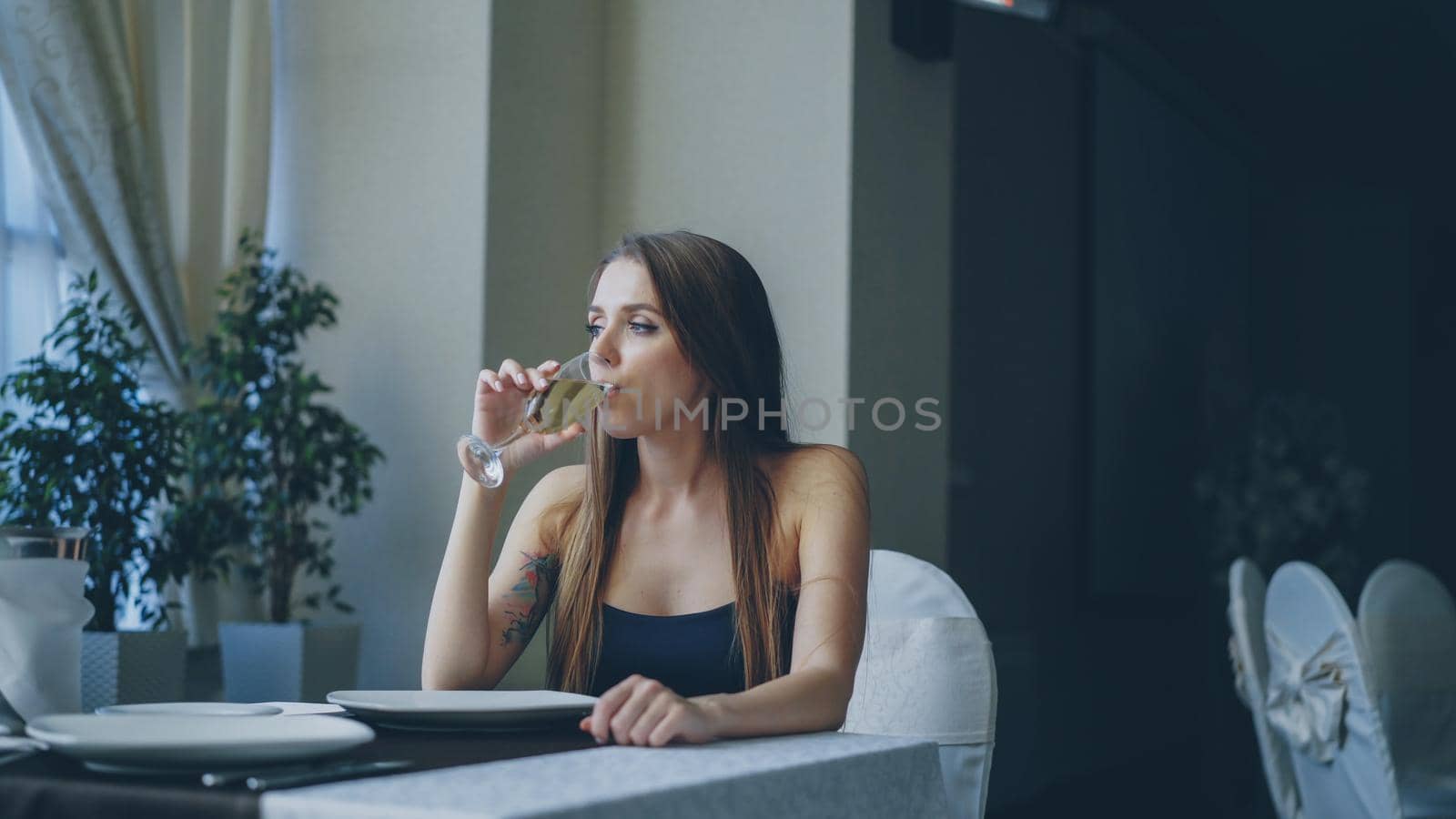 Pretty young woman is sitting alone at table in restaurant, drinking champagne and waiting for her boyfriend. Girl wearing beautiful dress is nervous and feeling uncomfortable.