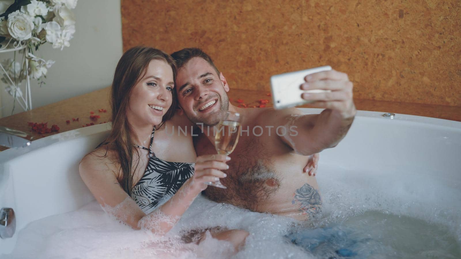 Pretty girl and her loving boyfriend are taking selfie with champagne glasses using smartphone while bathing in bathtub. They are smiling and posing looking at camera. by silverkblack