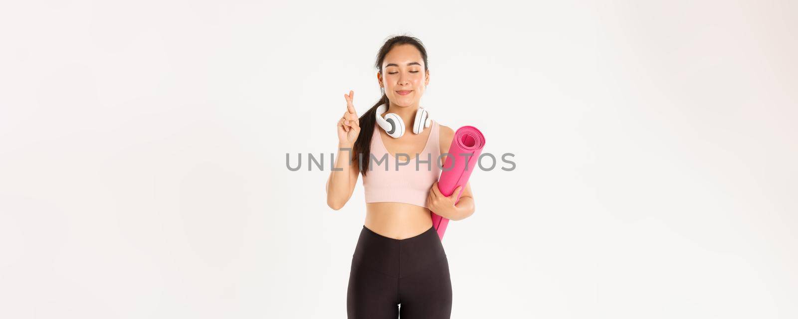 Sport, wellbeing and active lifestyle concept. Hopeful cute asian girl dreaming of going to gym after covid-19 quarantine, holding rubber mat and cross fingers while making wish.