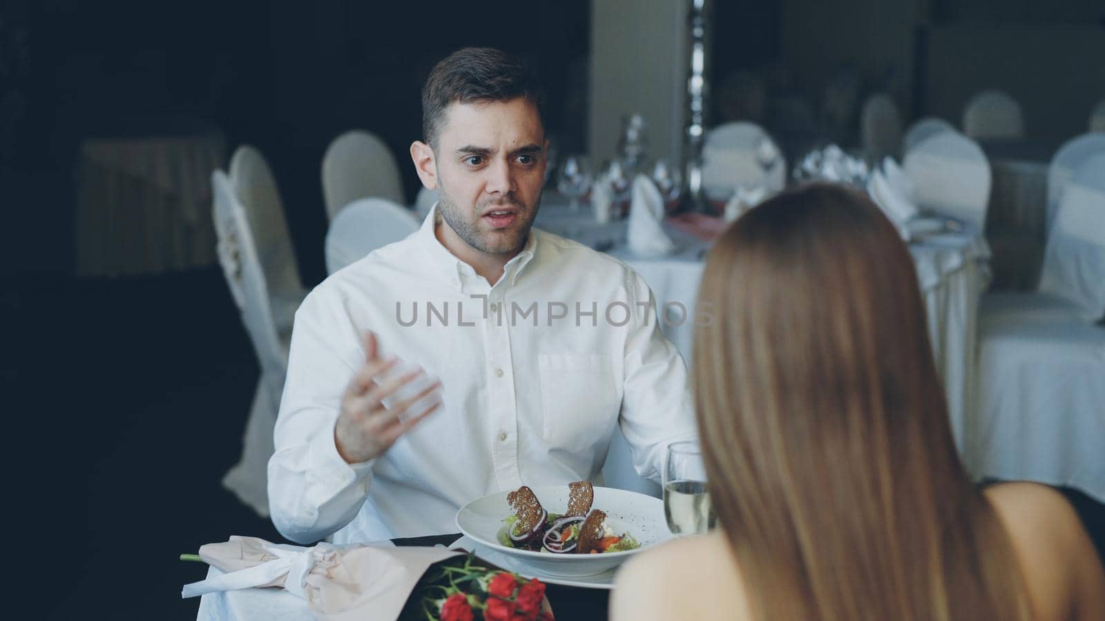 Young couple is quarreling while dining in restaurant, shouting and gesturing. Bouquet of roses, sparking champagne glasses and plates with food are visible. by silverkblack