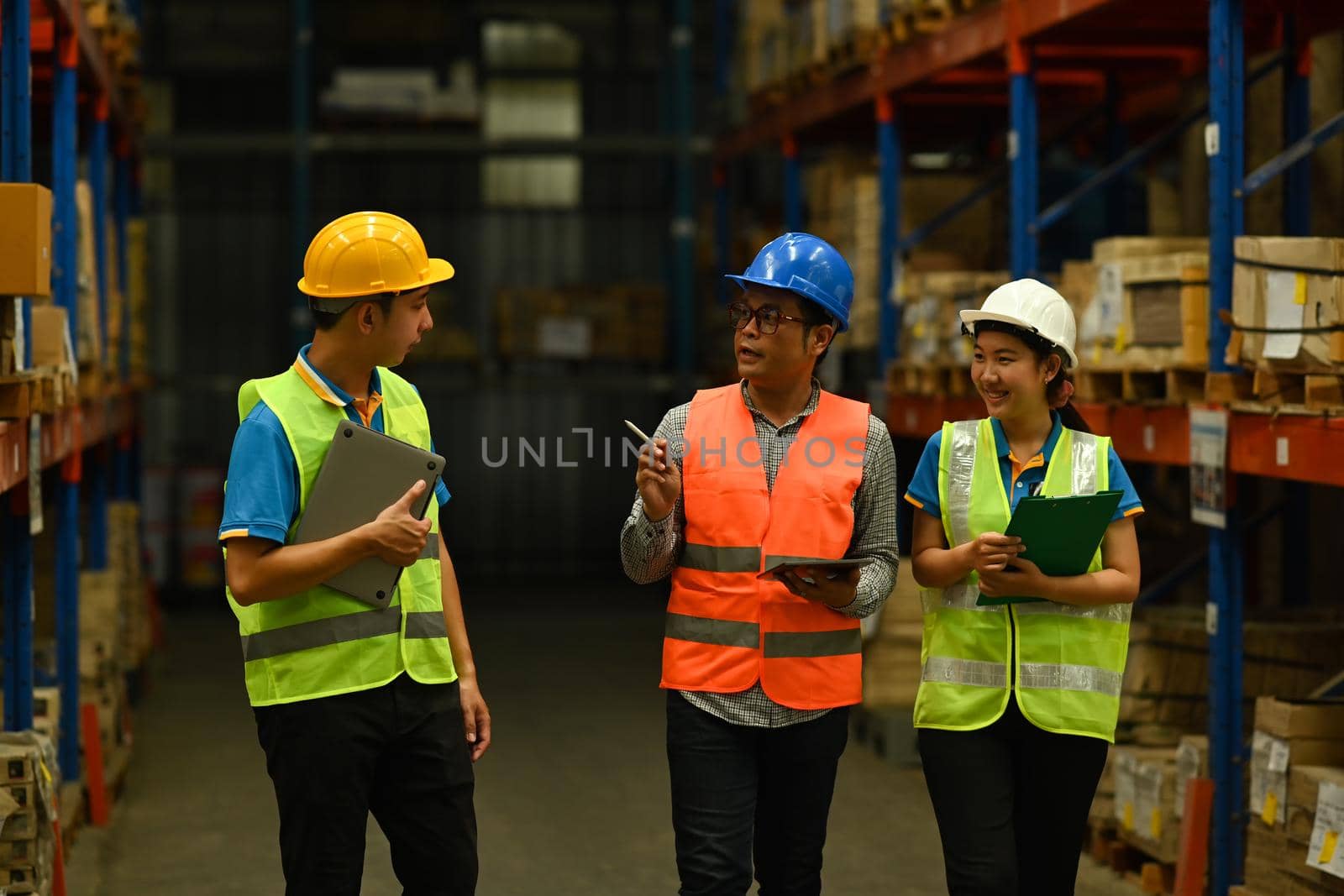 Mature male manager and workers in hardhats and reflective jackets checking quantity of storage product on shelf in a large warehouse by prathanchorruangsak