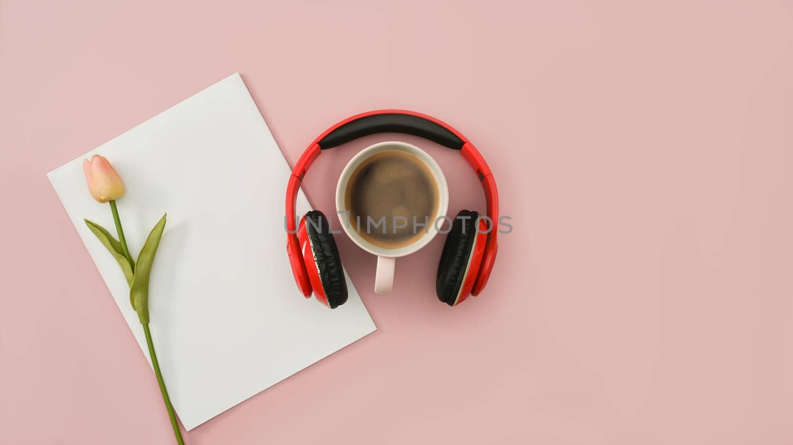 Red wireless headphones, coffee cup, blank paper and tulip on pink background. Top view, flat lay with copy space.